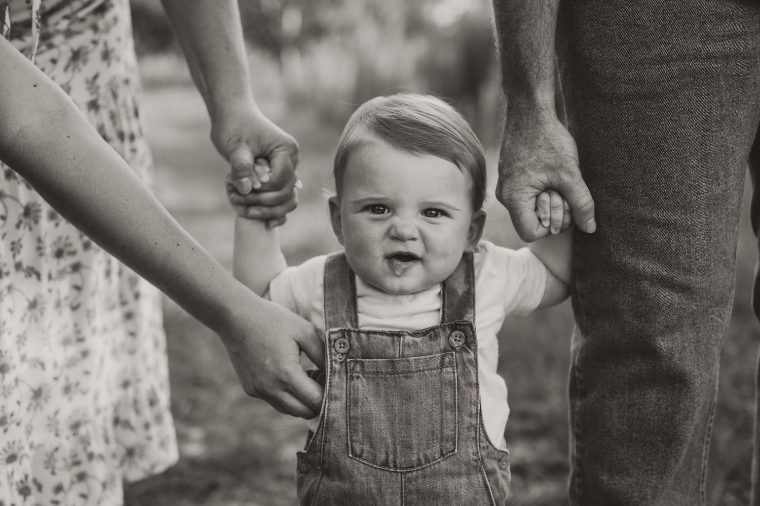 A toddler in denim overalls holds hands with parents in black and white photography.
