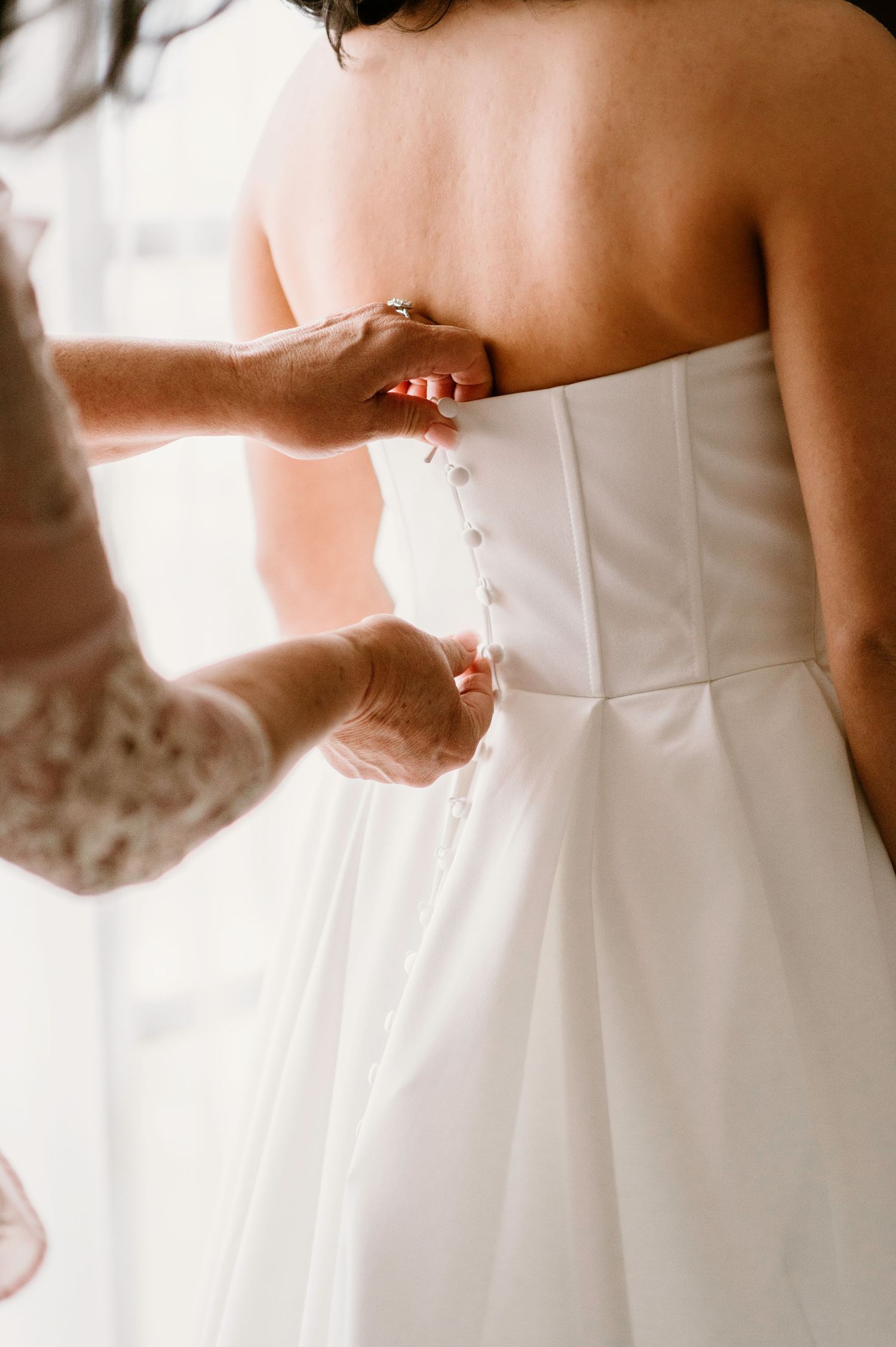 Detail shot of hands fastening the back buttons of a white wedding gown.