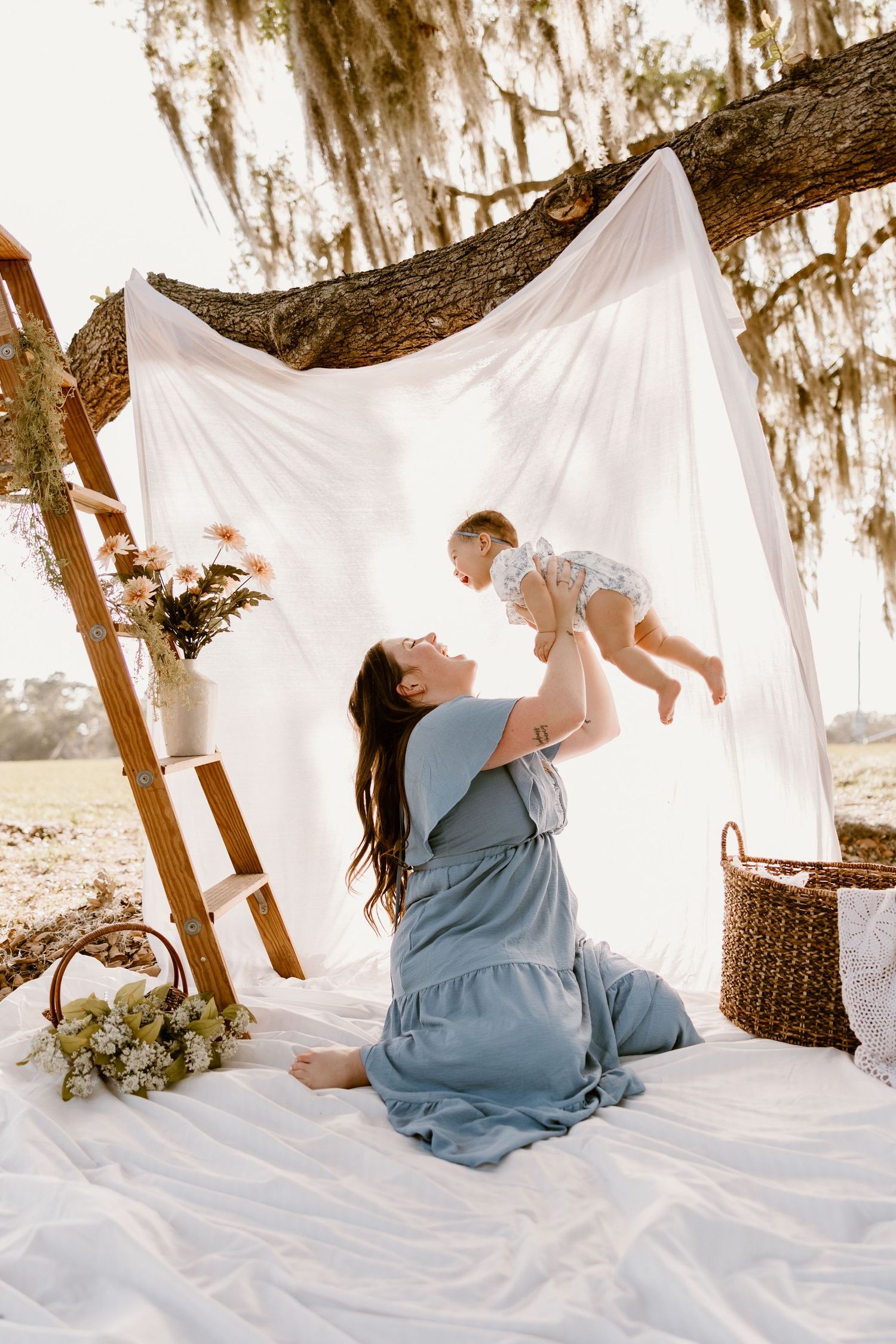 Dreamy outdoor photo session under Spanish moss with white fabric backdrop and rustic ladder against sunlit background.