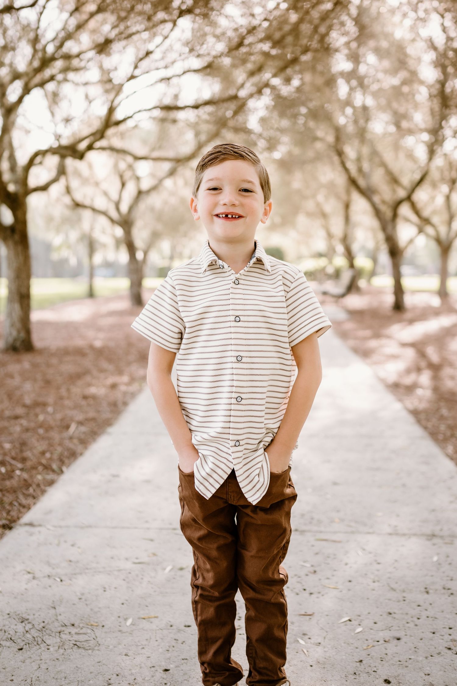 A child in a striped shirt and brown pants poses on a sunlit path.