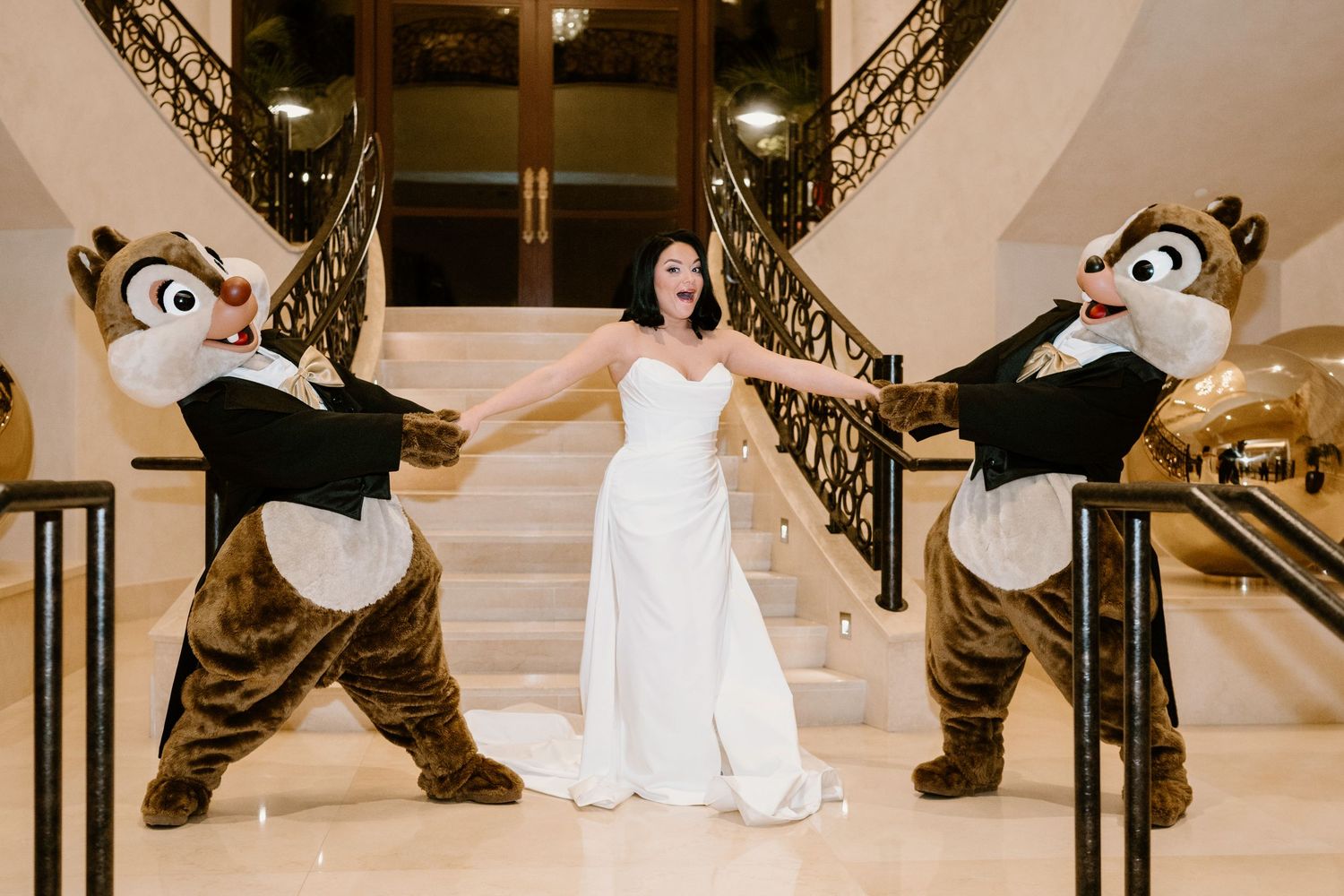A bride in a white gown poses with two Chip and Dale Disney character mascots on an elegant staircase.