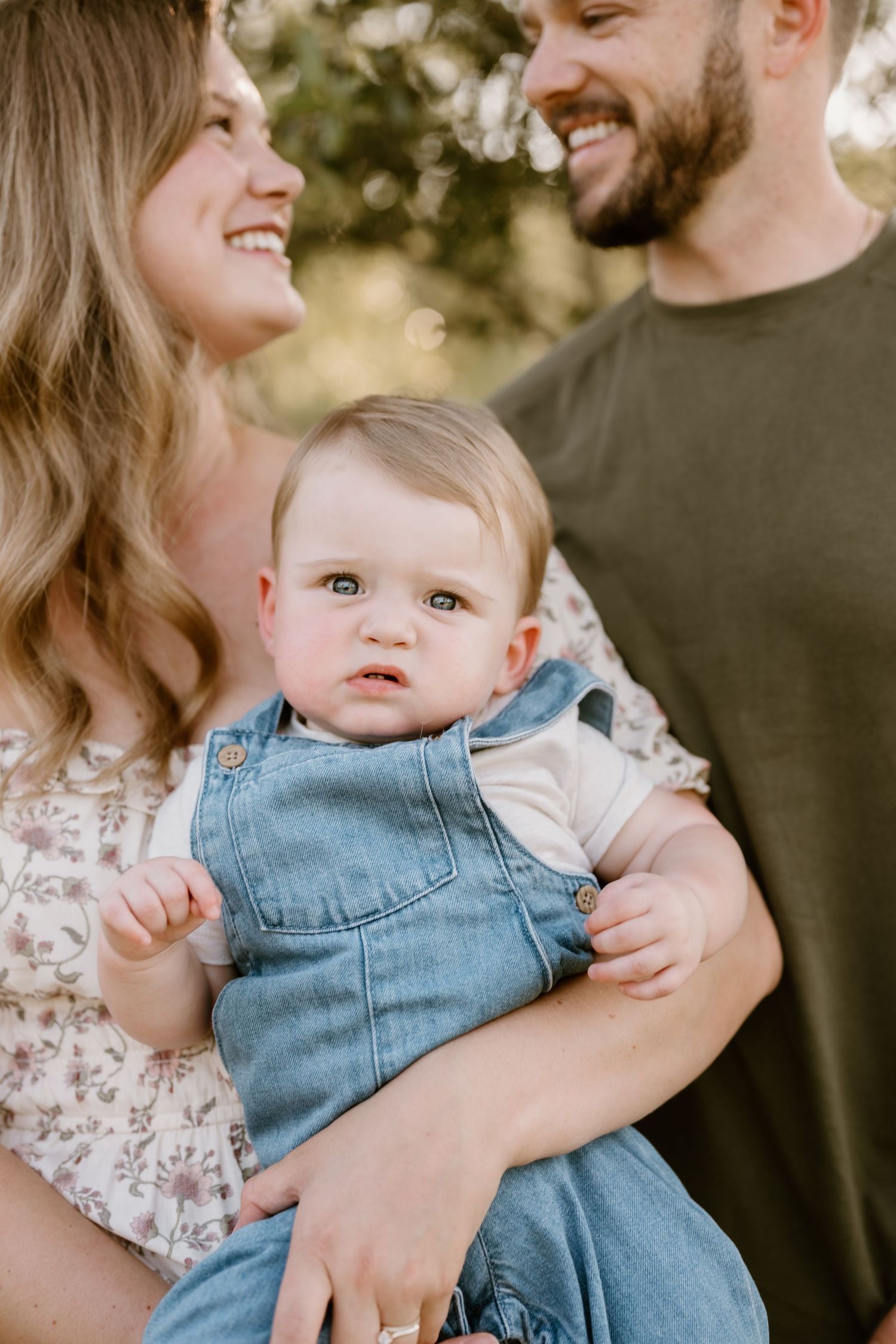 Baby in denim overalls being held and kissed by parents in casual attire.