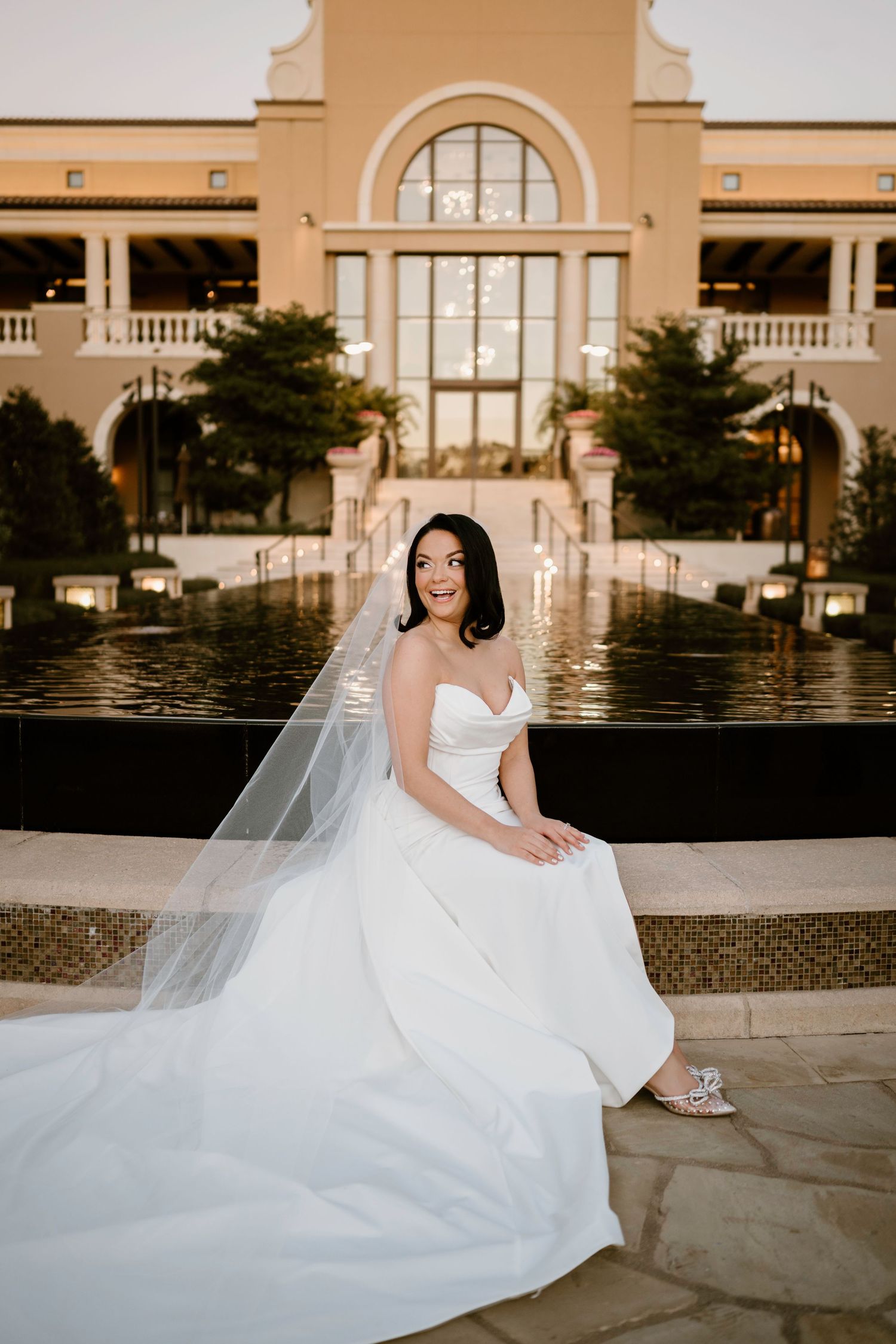 Bride in classic white gown sits gracefully by ornate resort fountain.