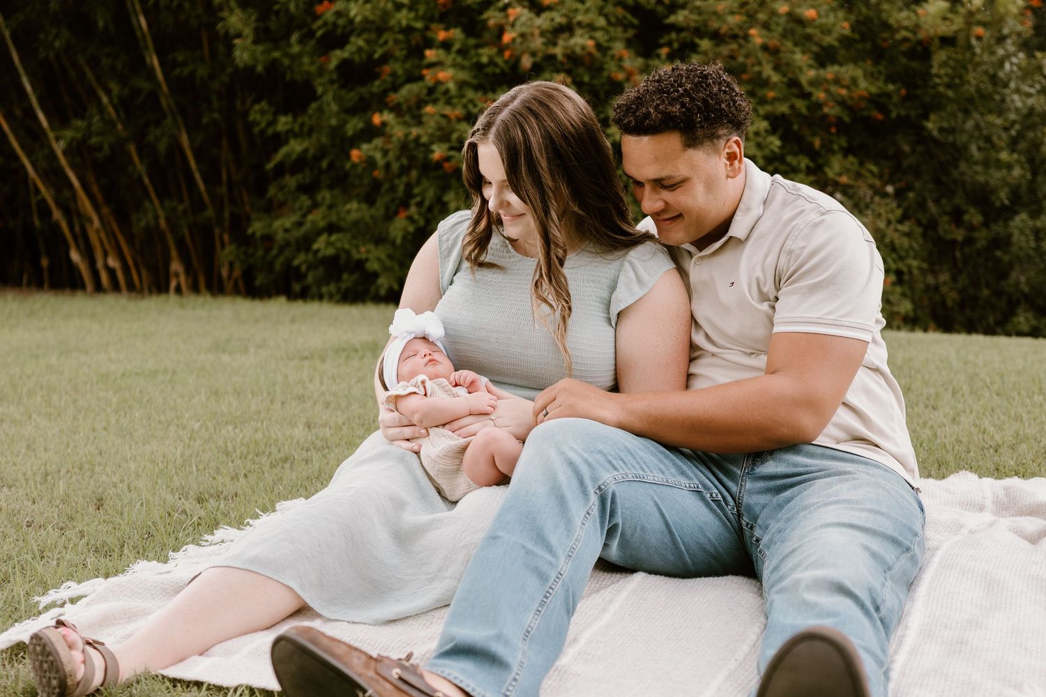 Family sits together on white blanket outdoors during lifestyle newborn photography session.