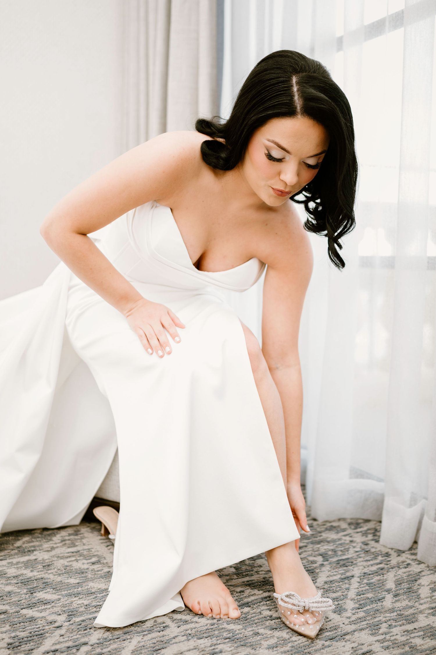 Bride in white dress adjusts her shoes in bright sunlit room.