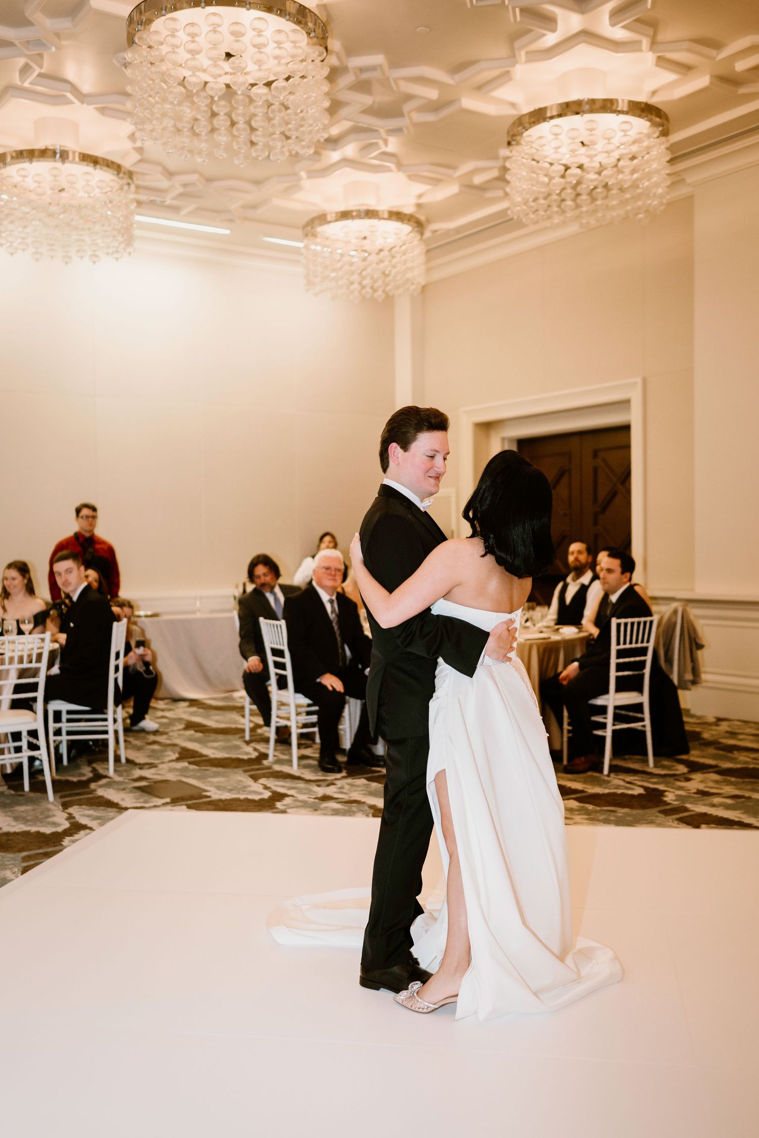 Couple performs first dance in an elegant ballroom with crystal chandeliers and white chairs.