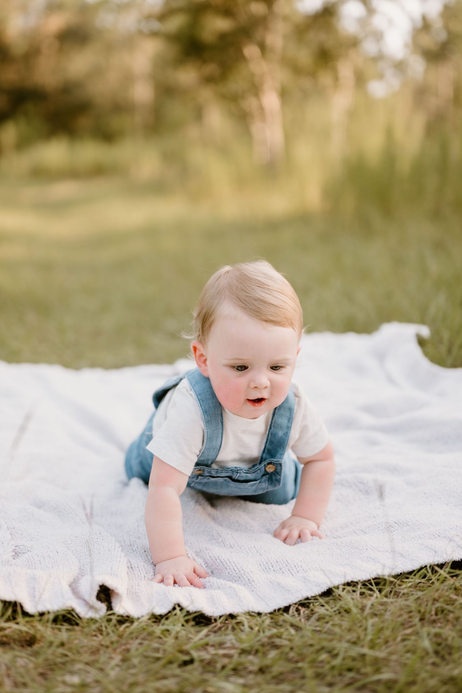 Baby in blue overalls crawls happily on white blanket during outdoor photo session.