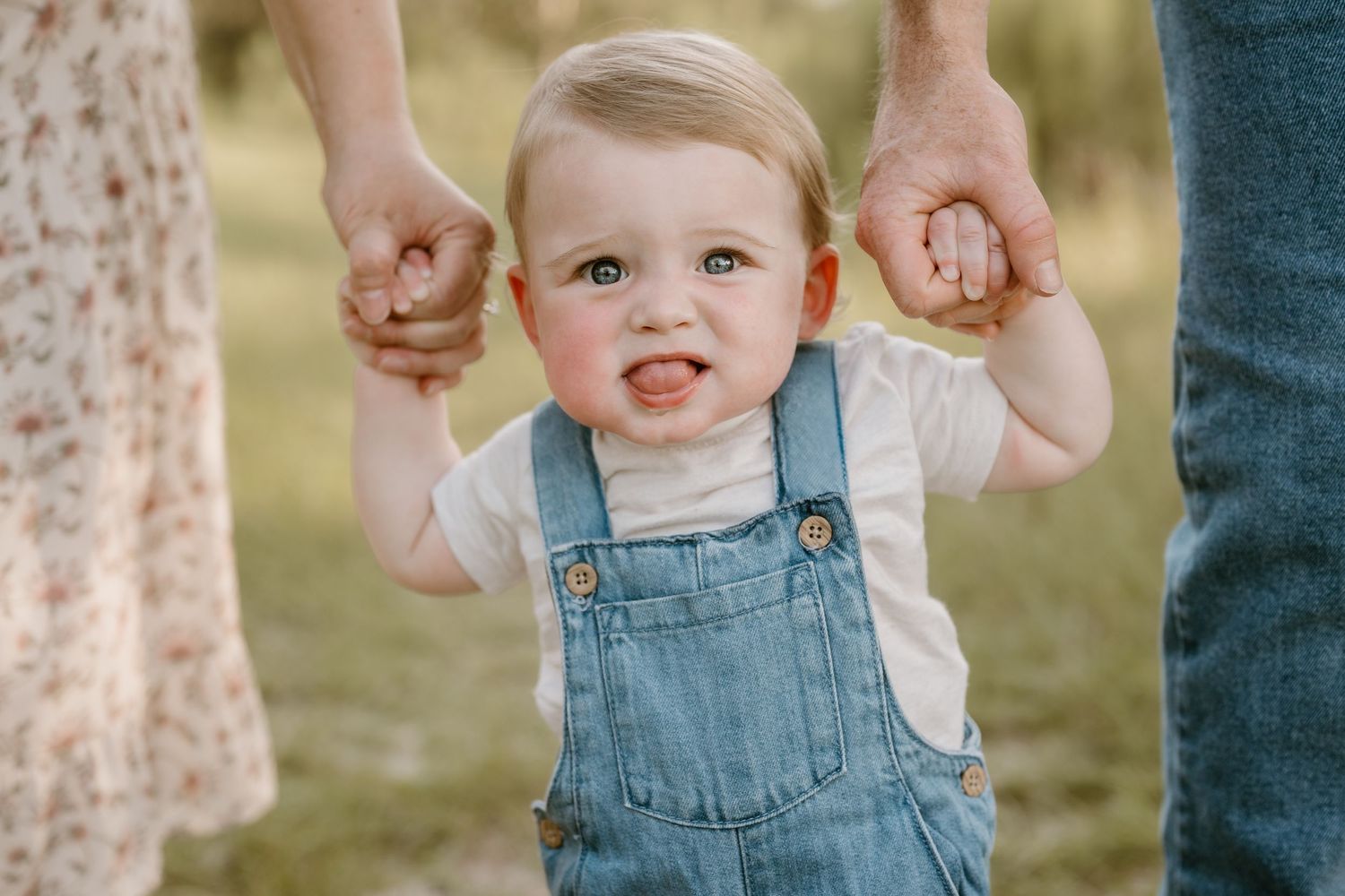 A toddler in denim overalls learning to walk while holding parents' hands in a sunny field.