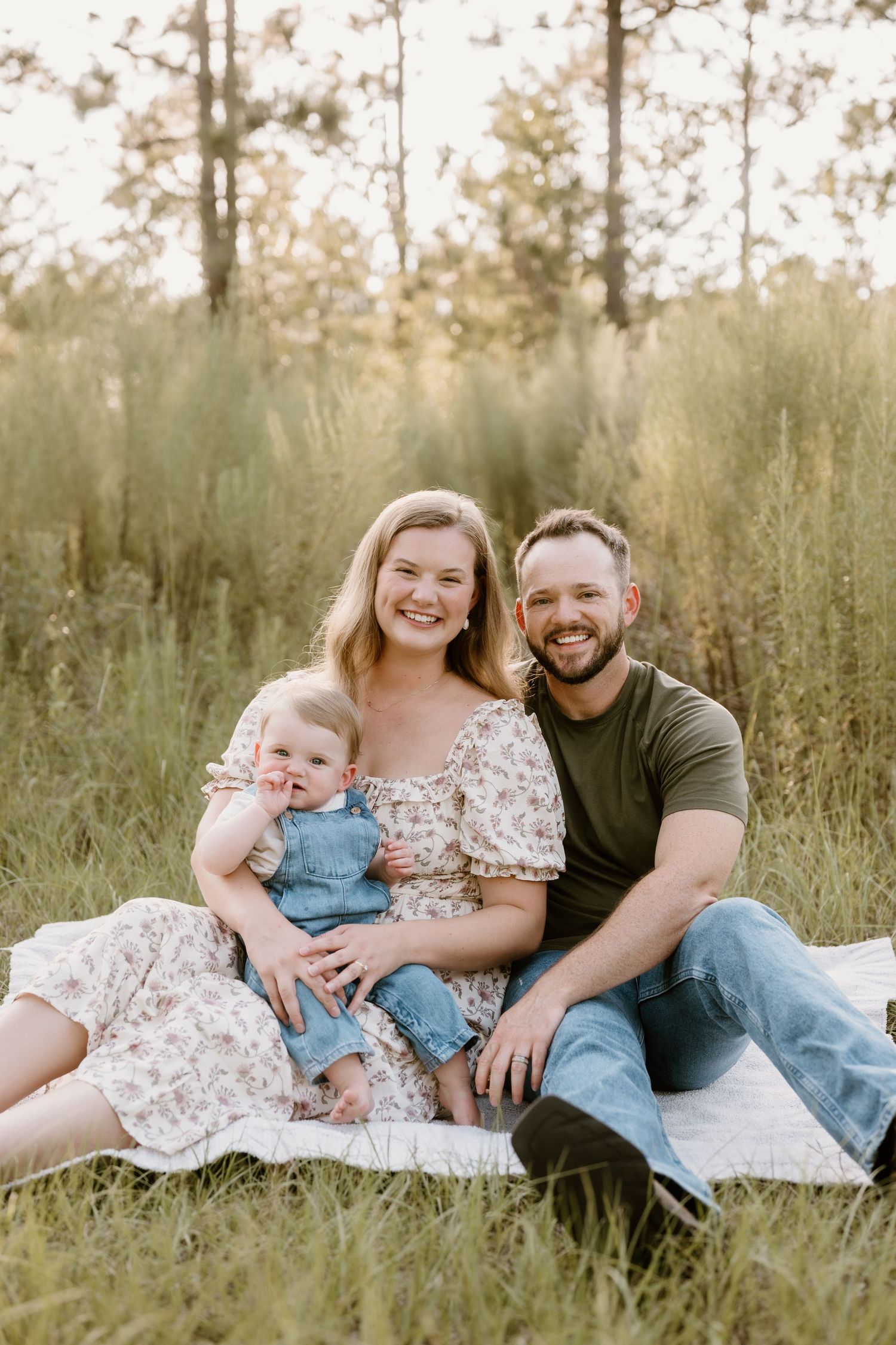 A family relaxes together on a white blanket in a sunny field with tall grass.