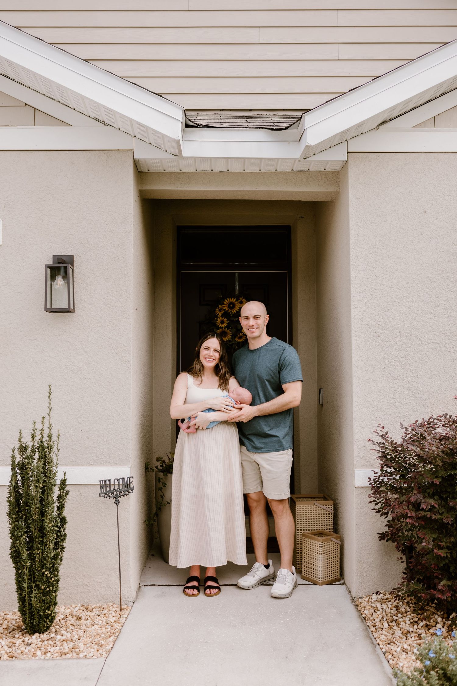 A couple poses with their newborn baby in front of a modern white house exterior.