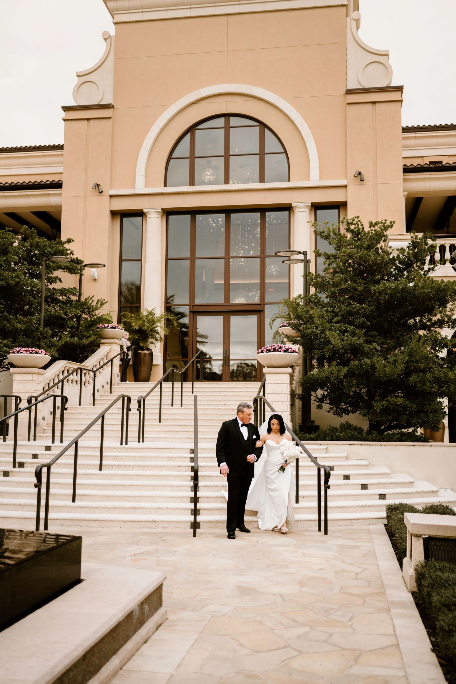 Wedding couple poses on stone steps of Mediterranean-style building with arched windows.