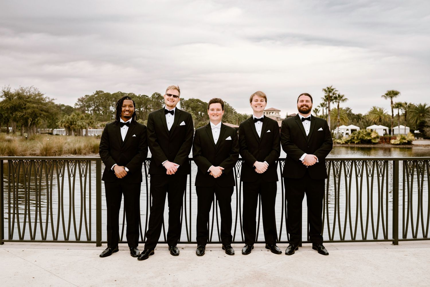 Groomsmen in black tuxedos standing in a line on a waterfront terrace.