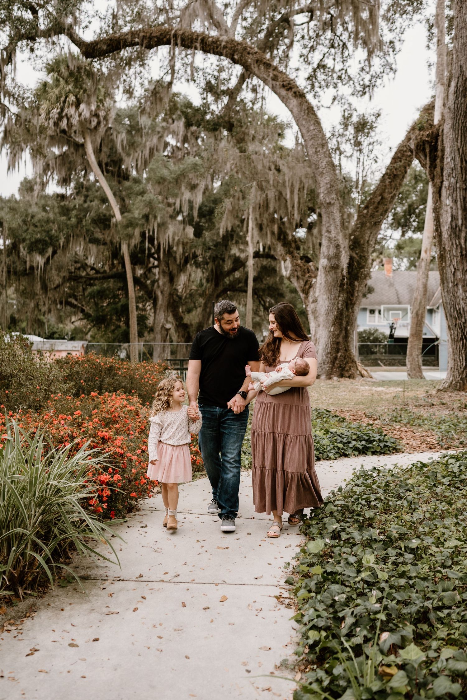 A family walks together on a garden path surrounded by Spanish moss draped trees and flowering bushes in a natural setting.