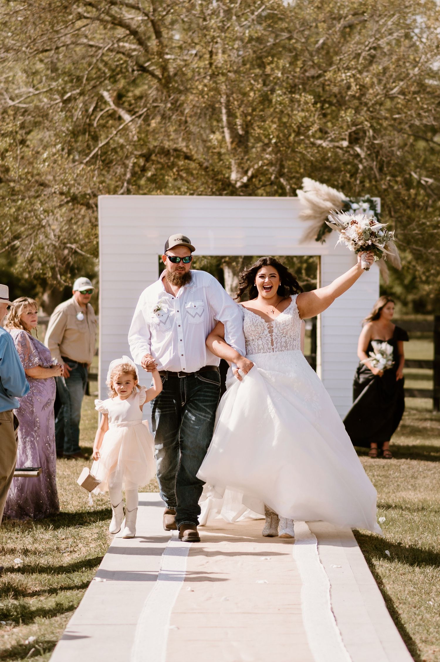 Newlyweds walk down an outdoor aisle after their ceremony with a flower girl following behind.