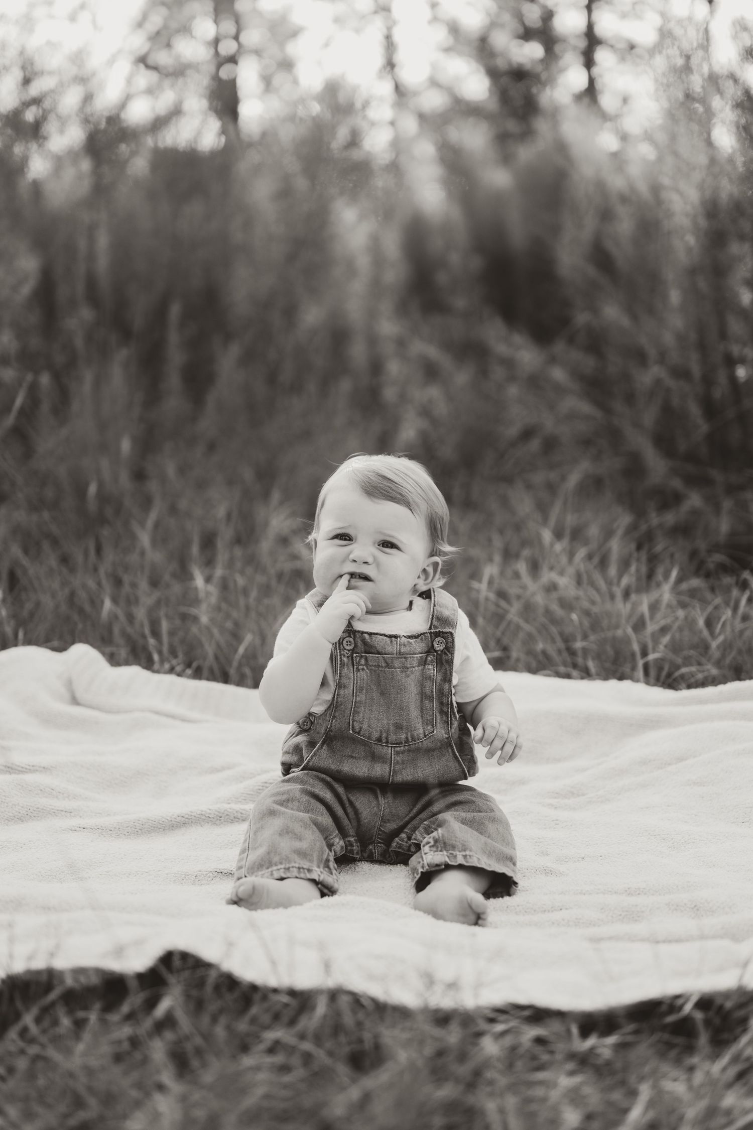 Baby wearing denim overalls laughs while sitting on white blanket during outdoor photo shoot.