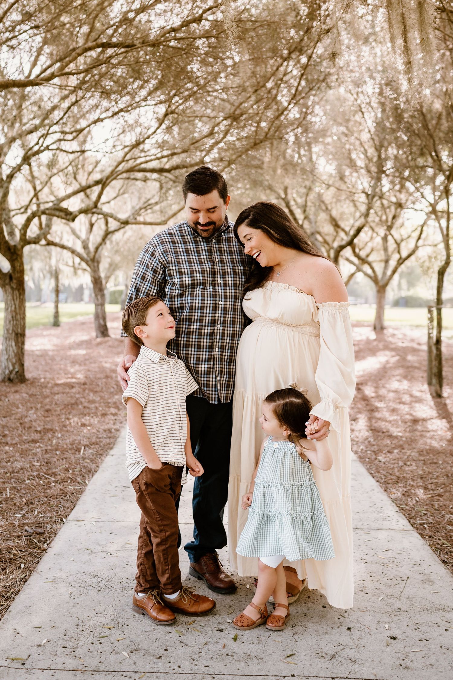 A family poses together on a tree-lined path in coordinated neutral outfits.
