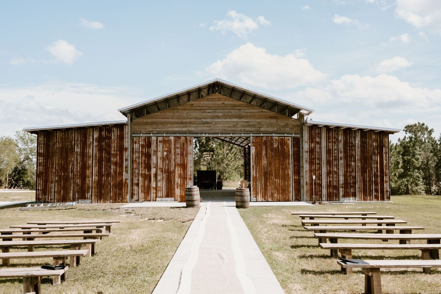 Rustic outdoor wedding venue with wooden benches and barn in background.
