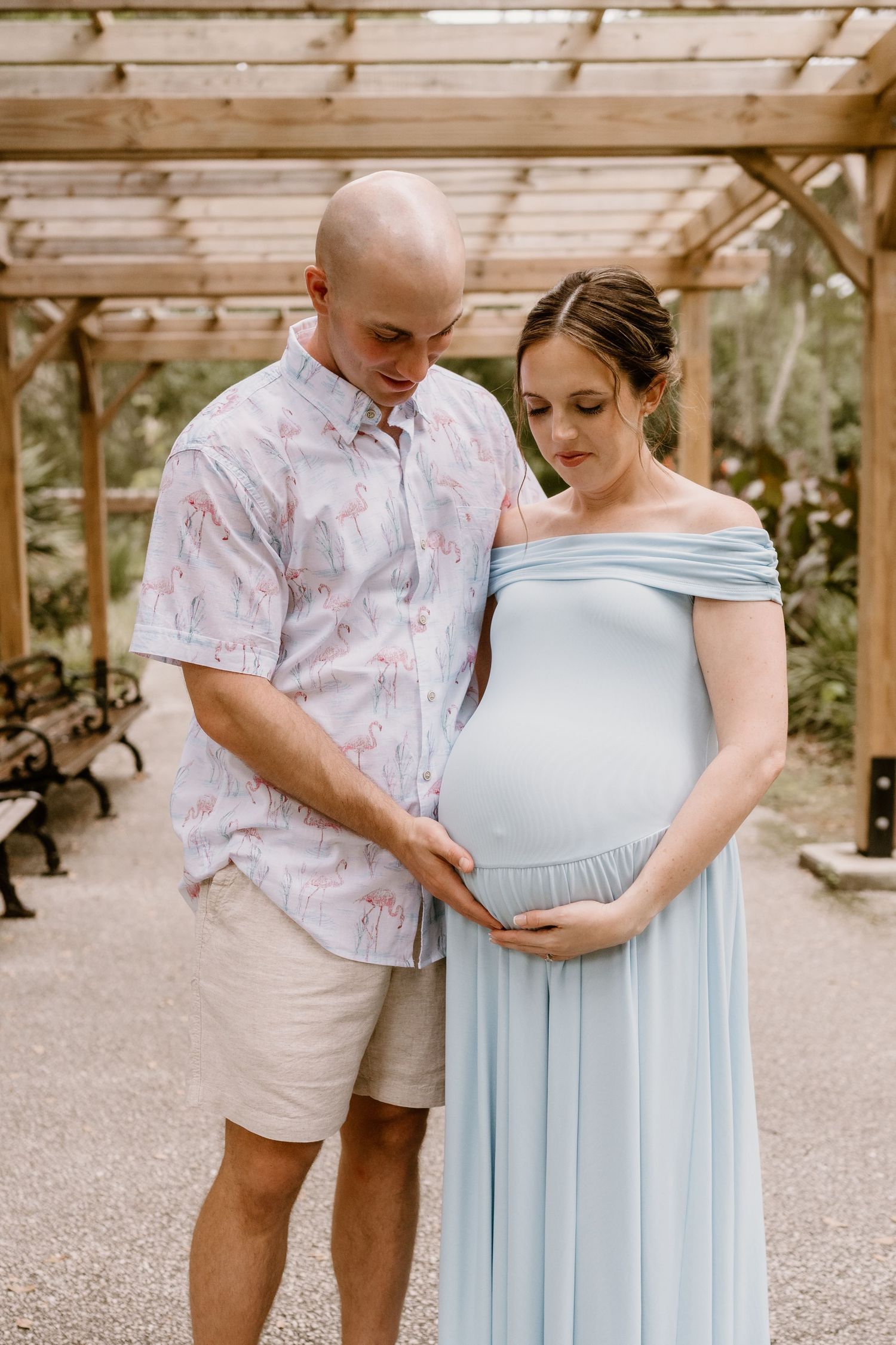 A couple in casual attire poses under a wooden pergola in a garden setting.
