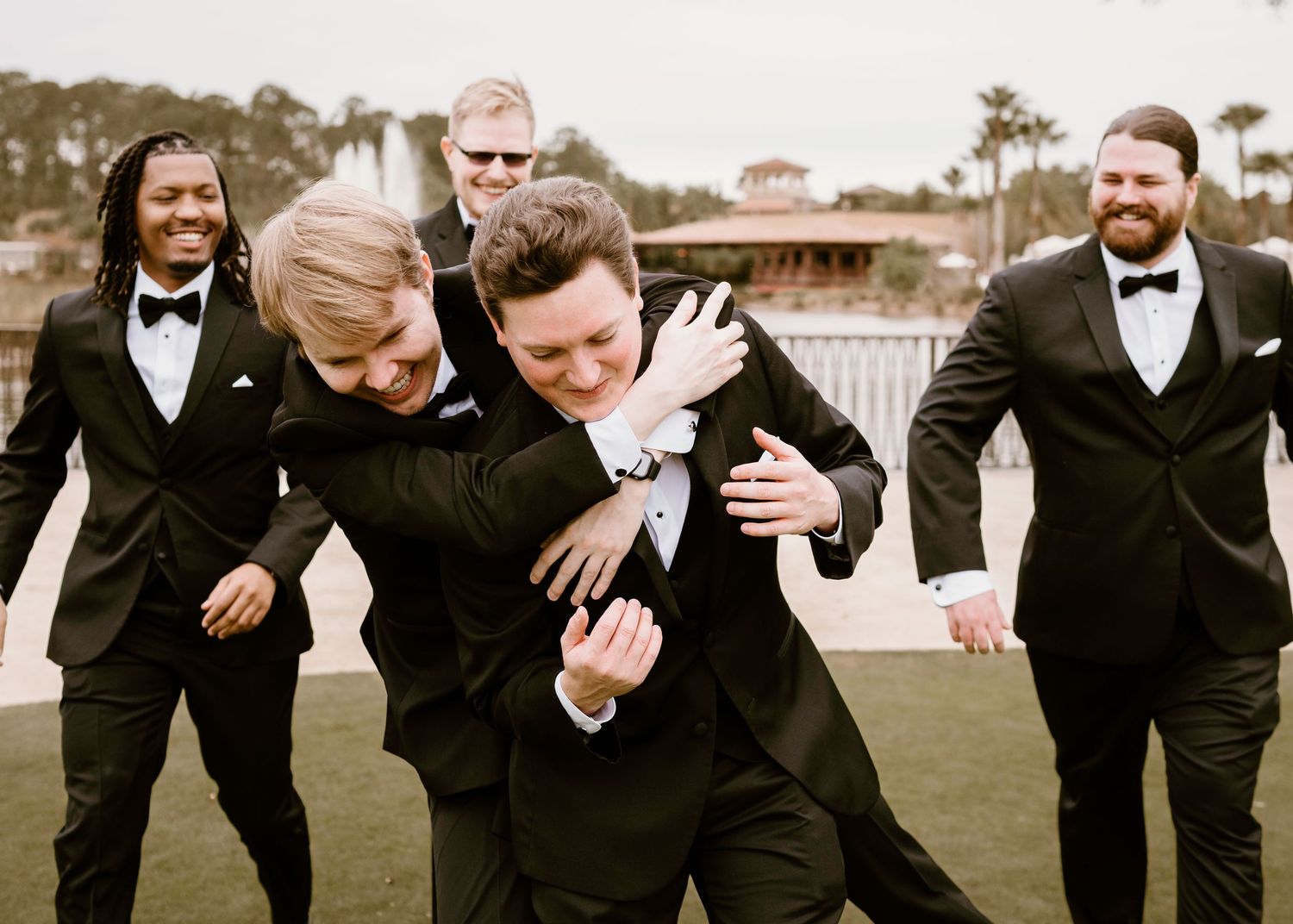 Playful groomsmen in black tuxedos sharing a candid moment outdoors.