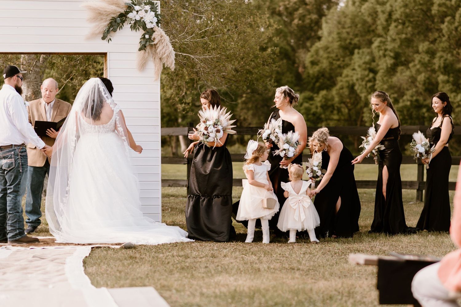 Wedding party poses outdoors with flower girls in white dresses and bridesmaids in black gowns.
