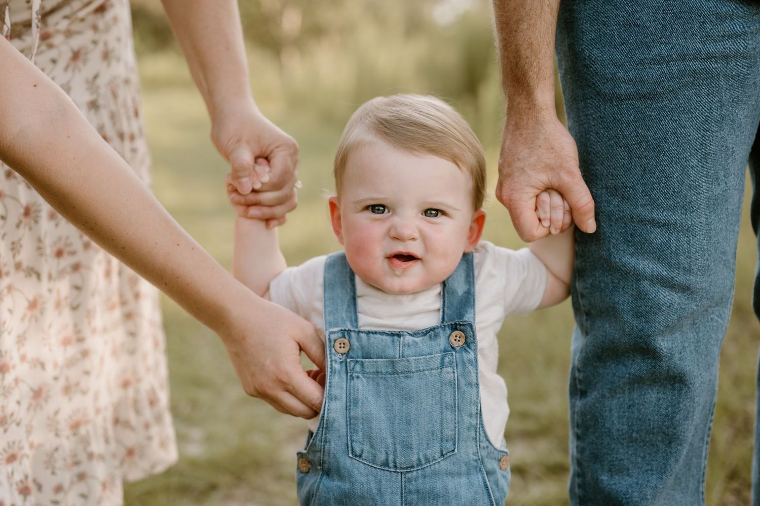 A small child in denim overalls holds hands with parents while learning to walk outdoors.