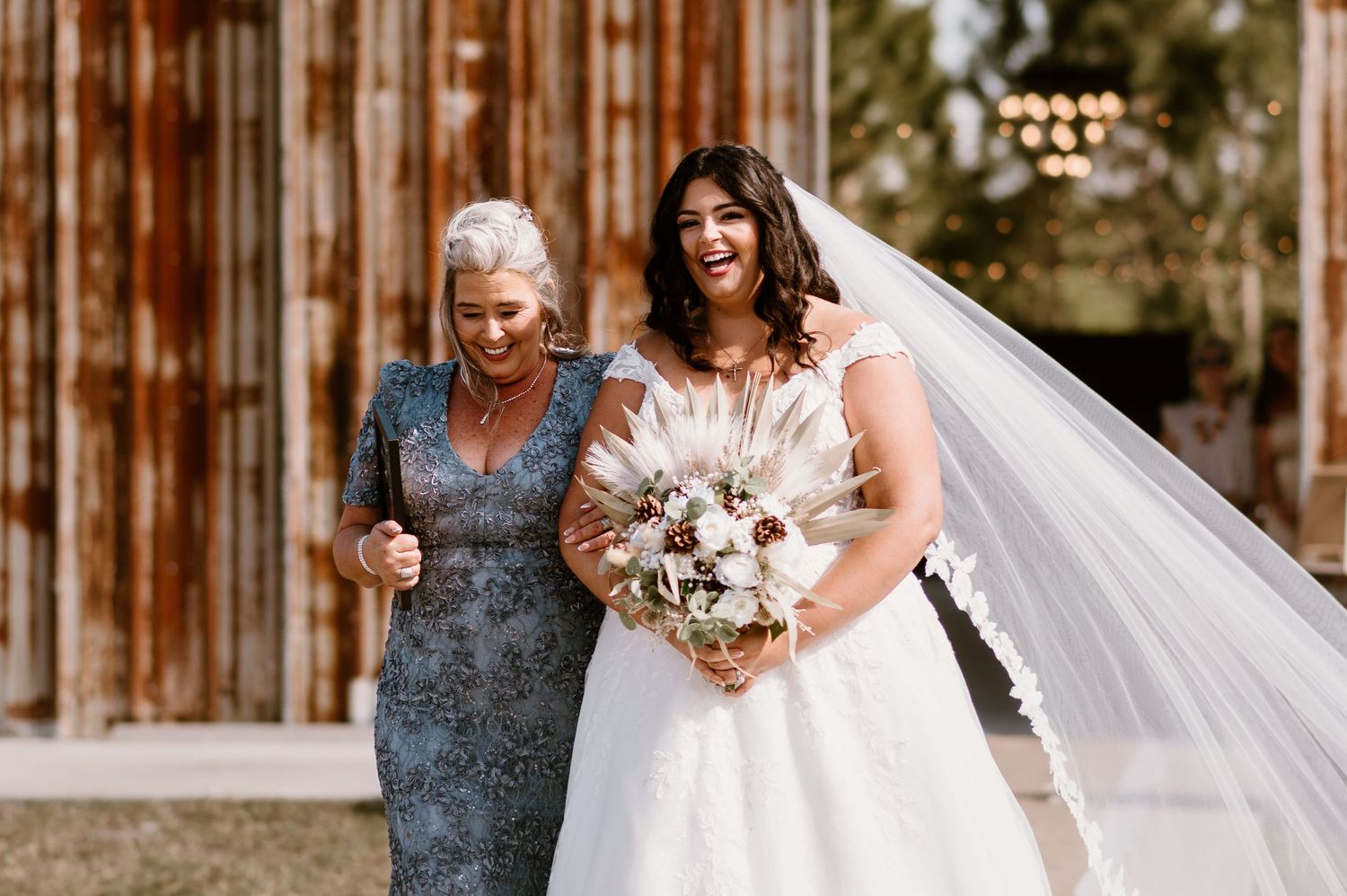 A bride in a white gown stands with an older woman in a gray dress holding bouquets at a rustic barn venue.
