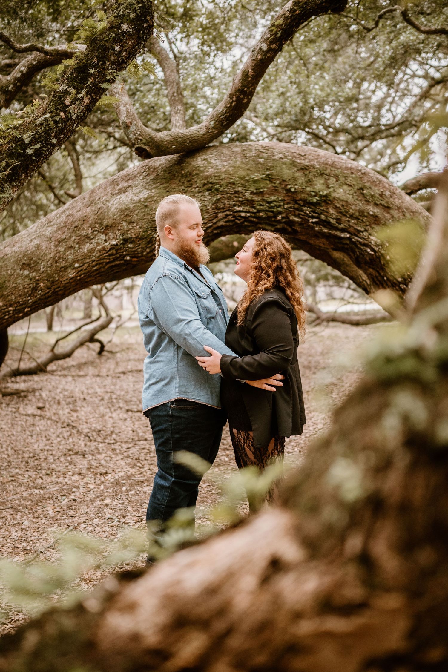 Romantic couple embracing under large oak trees in a natural setting.
