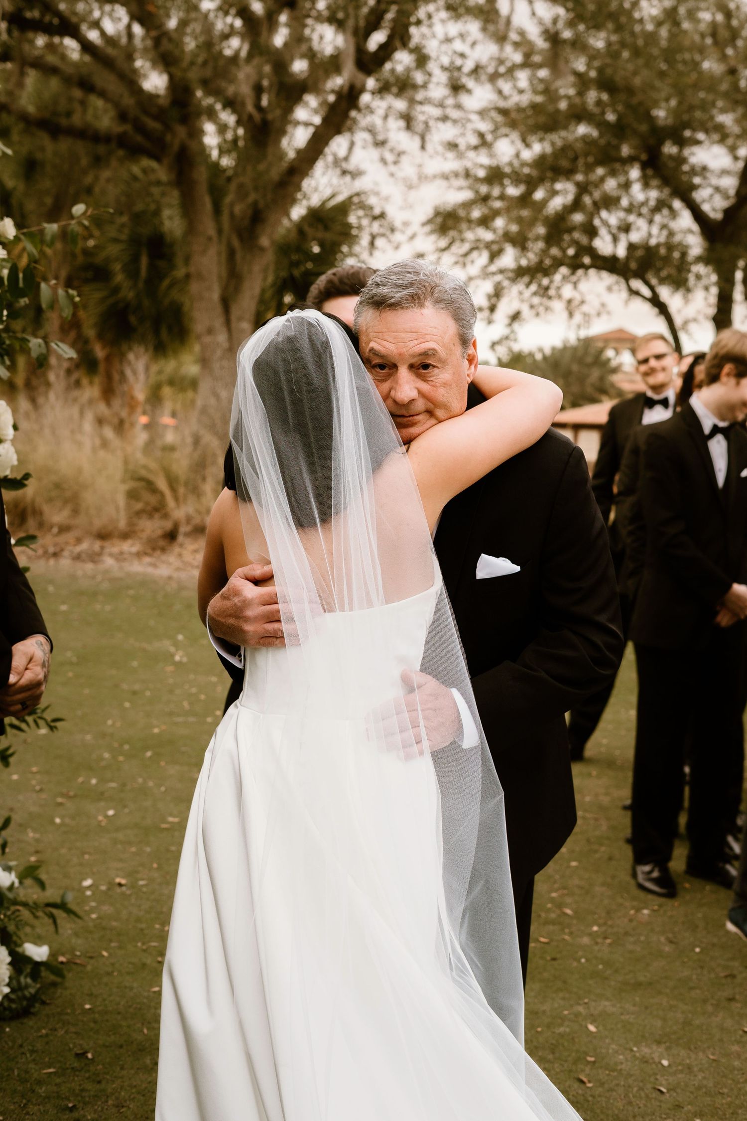 A heartfelt embrace between a bride in white and someone in formal attire during an outdoor wedding ceremony.