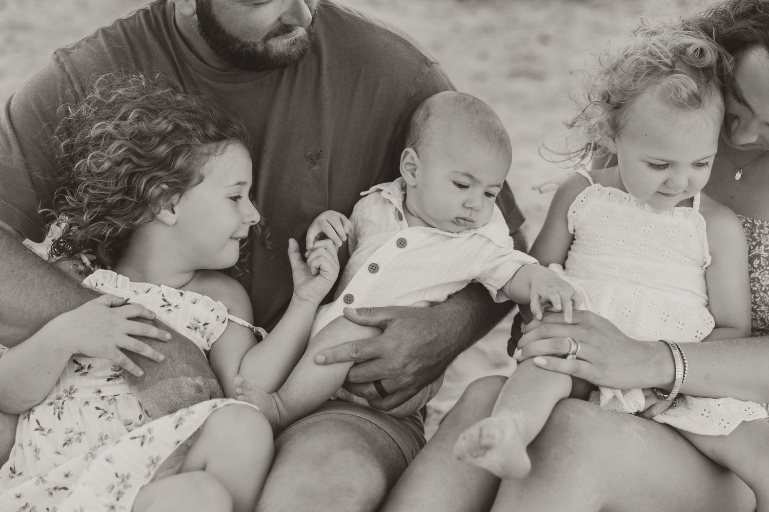 Black and white family photo with father holding newborn baby surrounded by two young daughters on a beach.