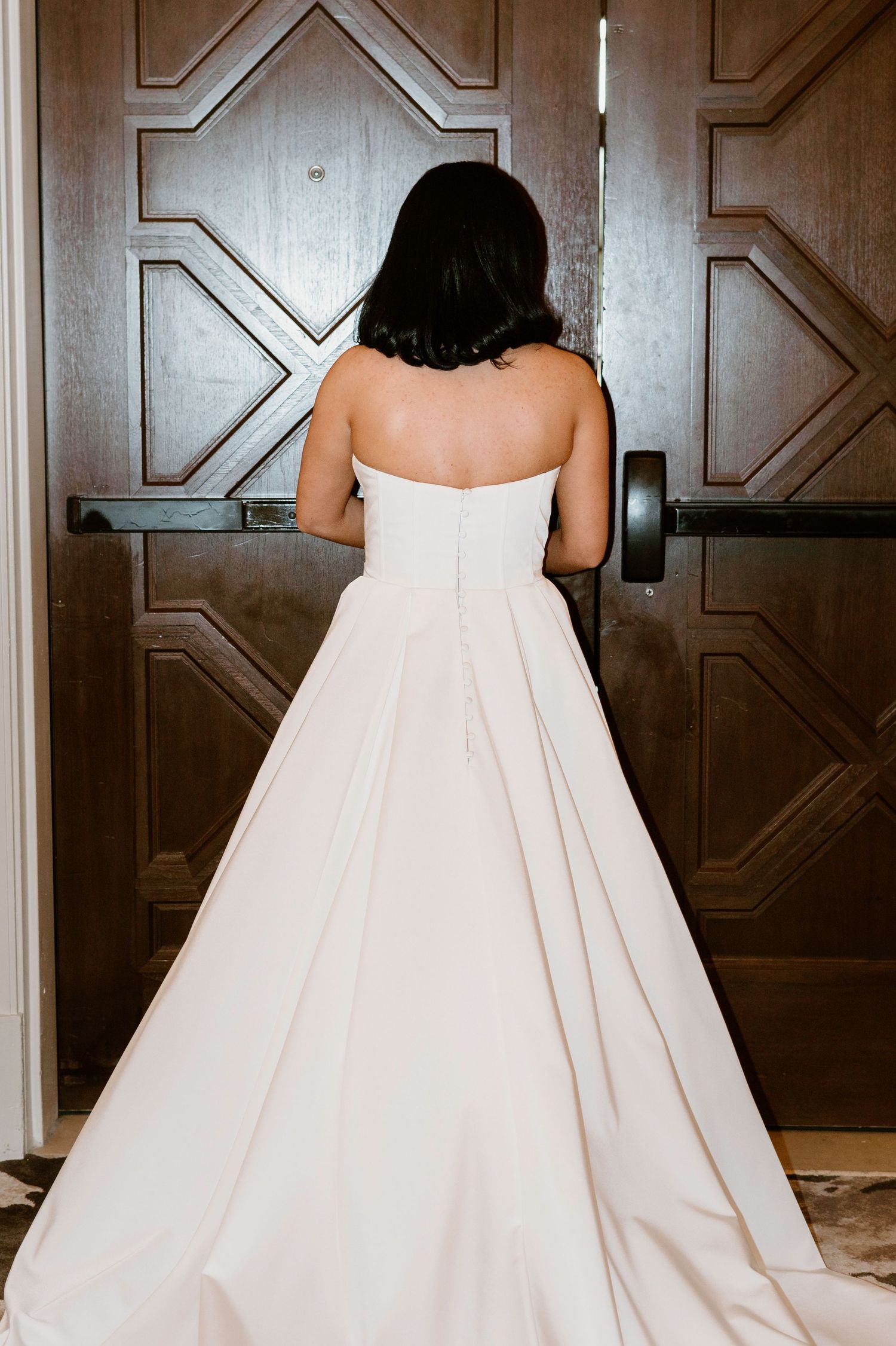 Back view of elegant white wedding gown against dark wooden paneled wall.