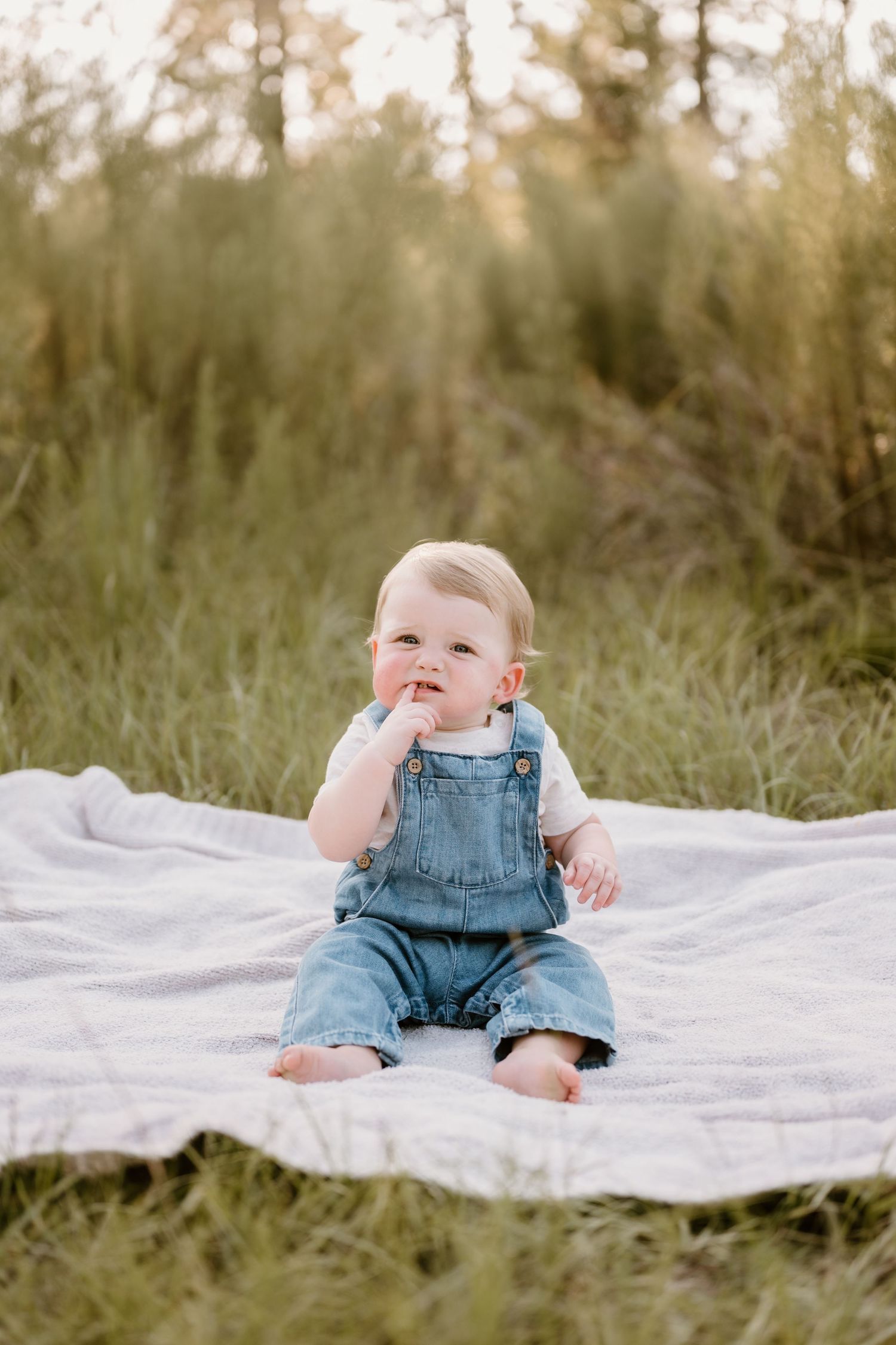 Baby wearing denim overalls sits and smiles on white blanket outdoors during golden hour photo session.