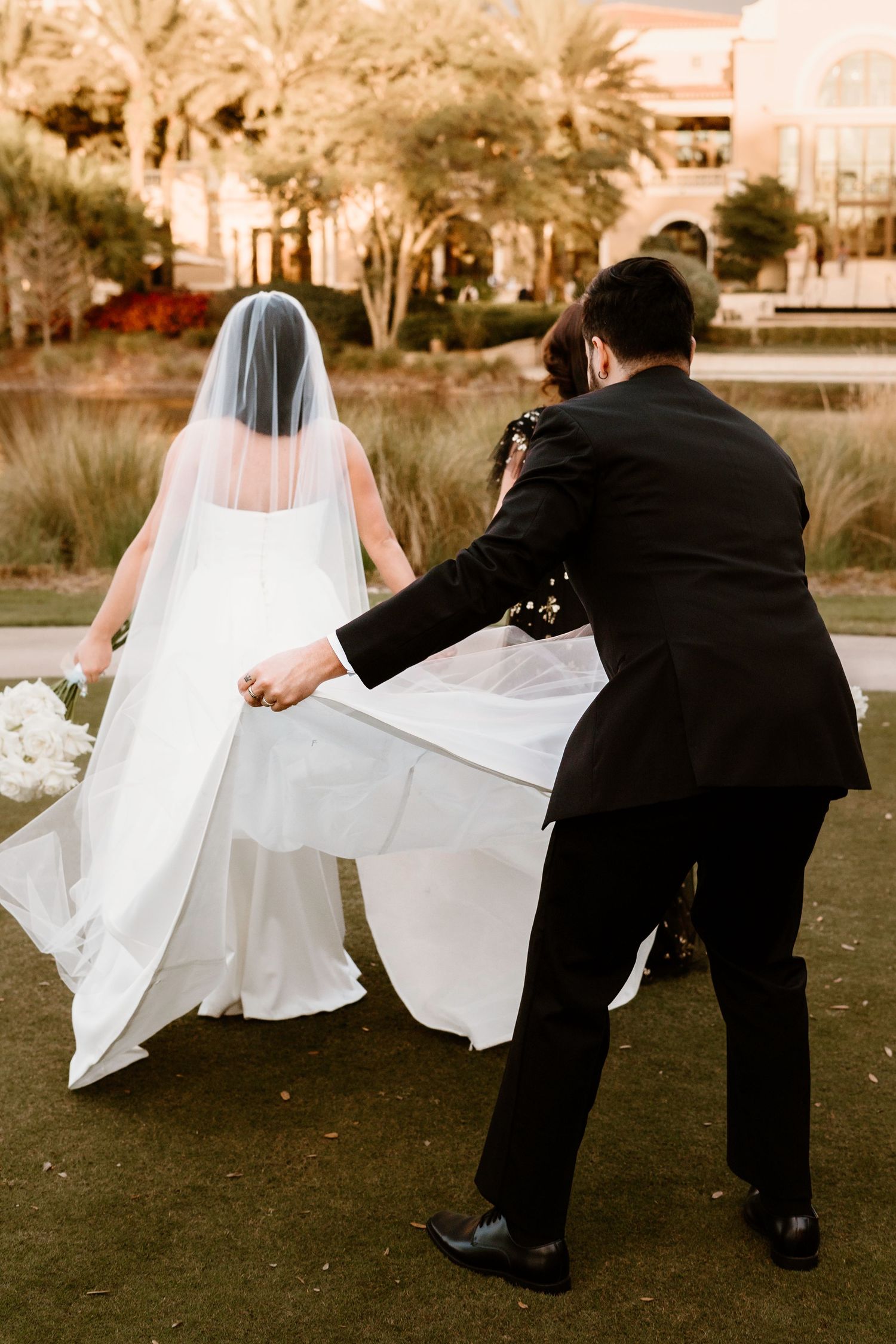 Wedding party members playfully helping with the bride's dress train on grass.