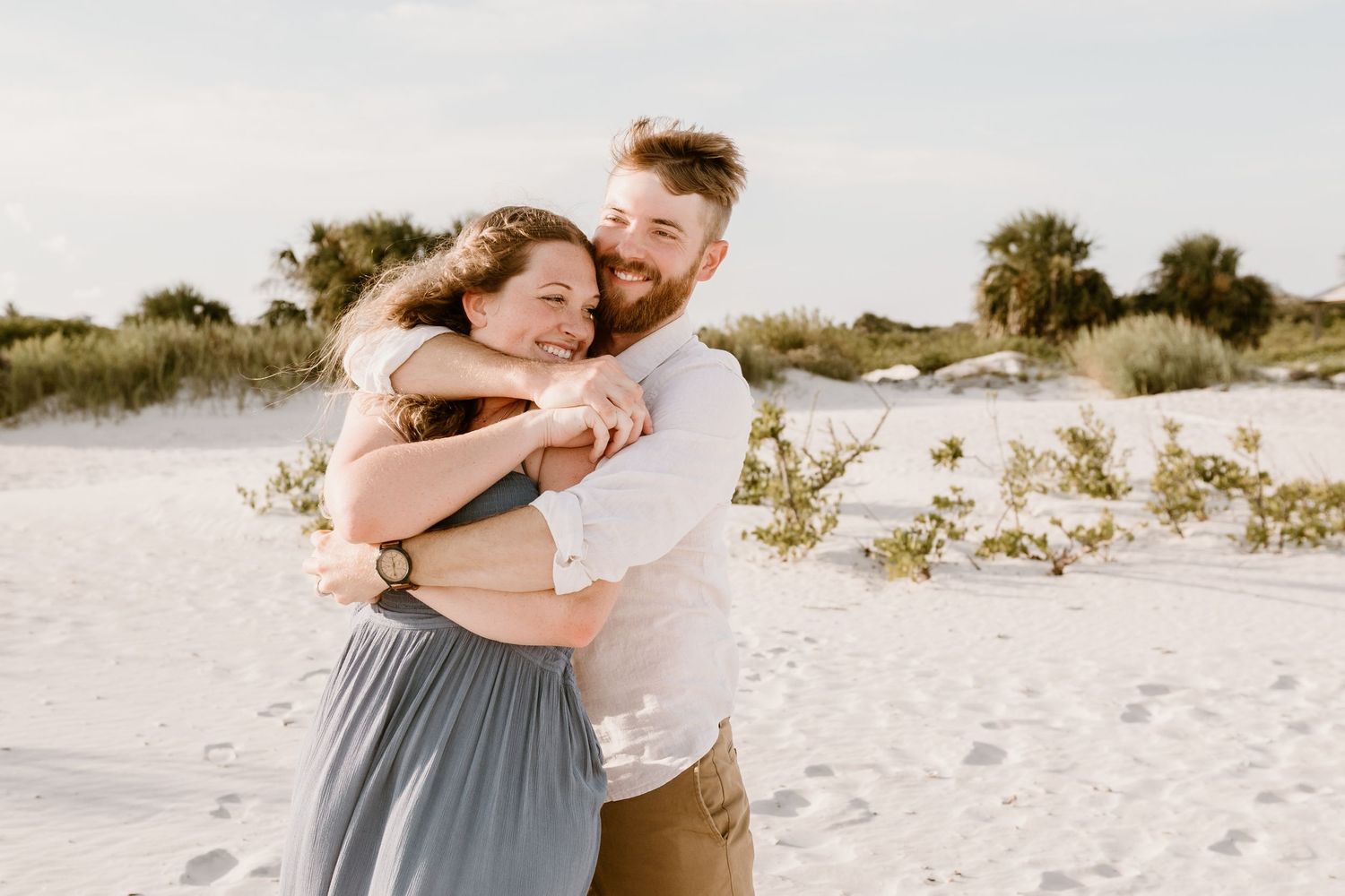 A couple shares a tender moment while dancing together on a white sandy beach at sunset.