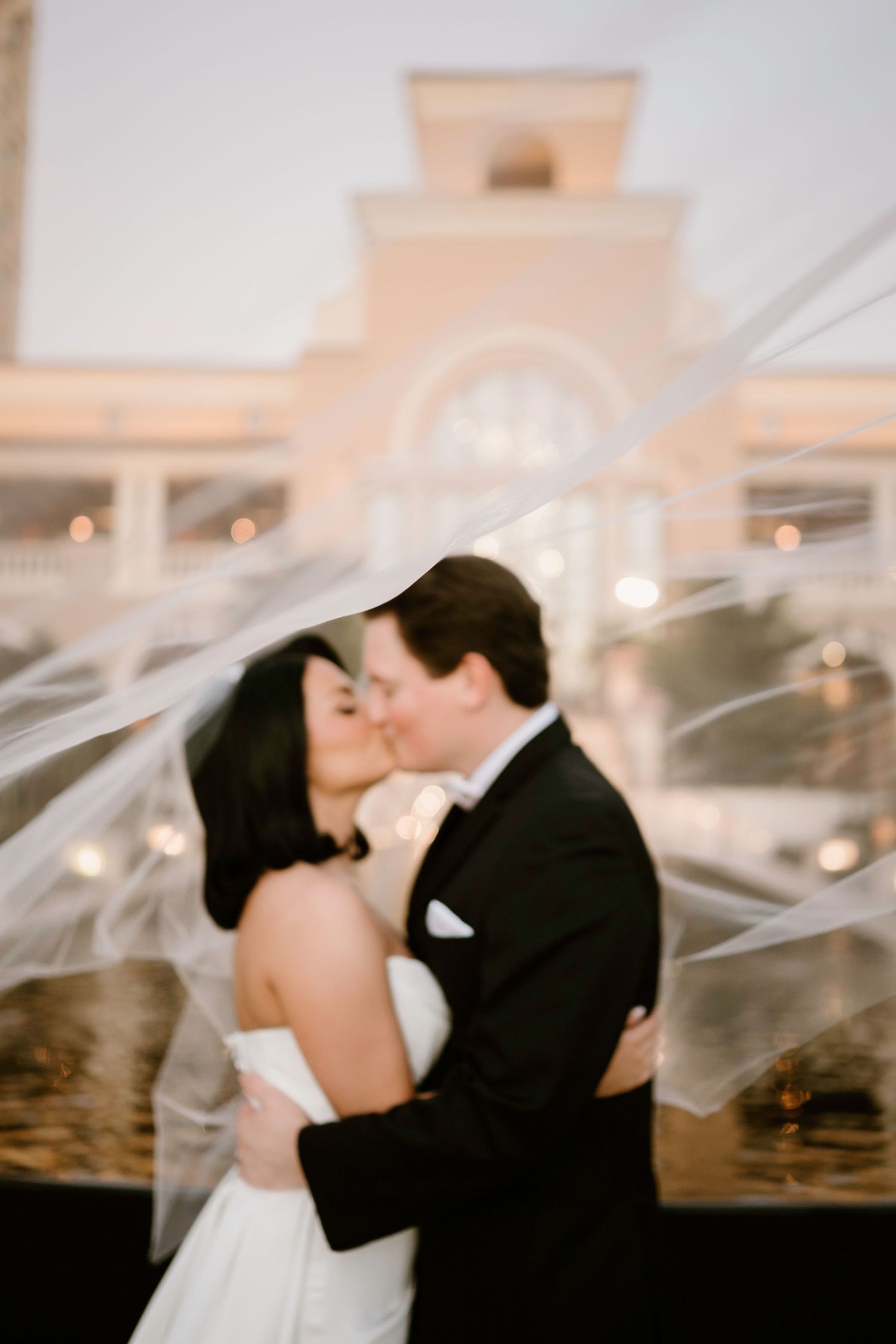 A romantic wedding couple shares a kiss under a flowing veil during sunset.