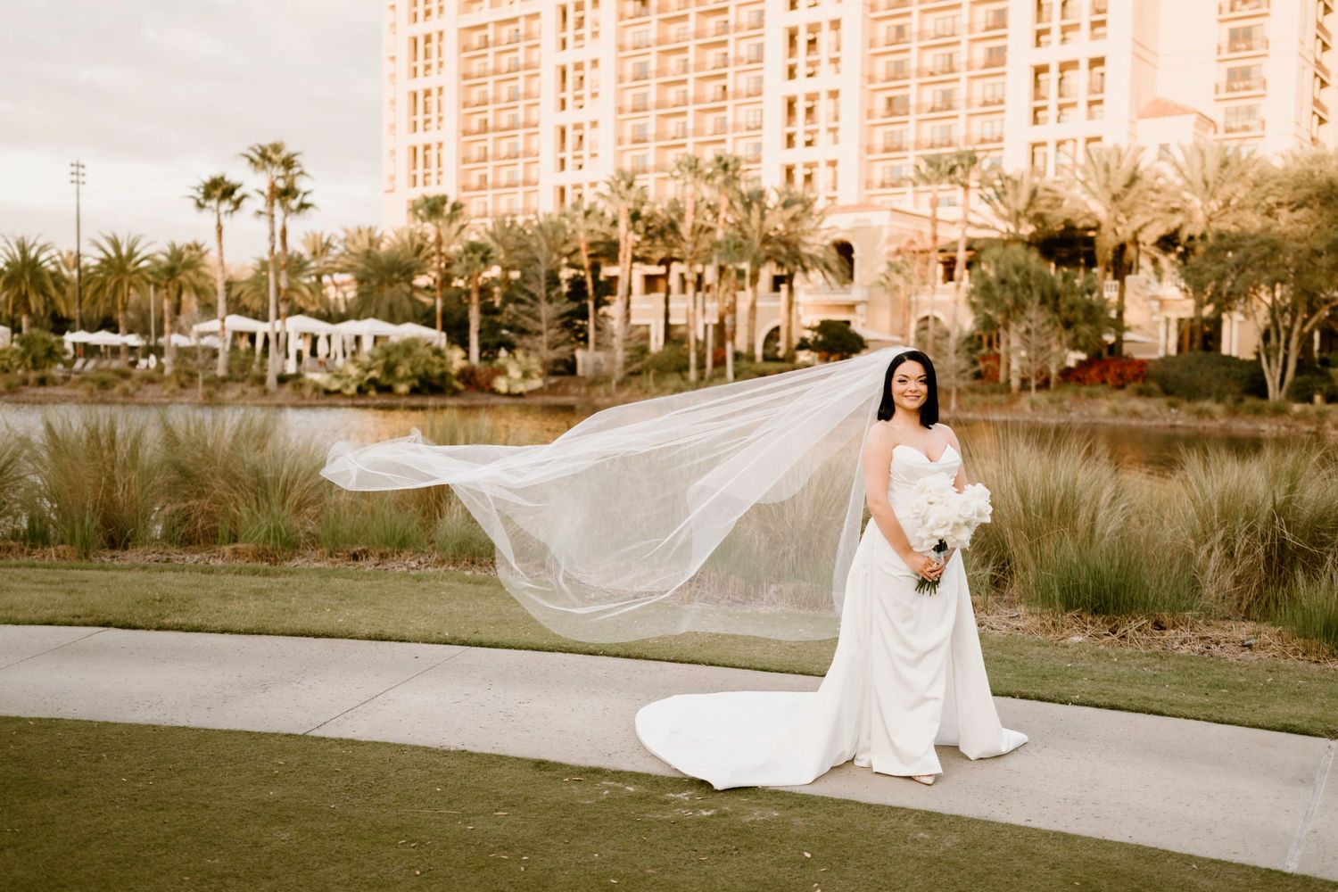 A bride in a flowing white dress poses on grass with a large building in the background.