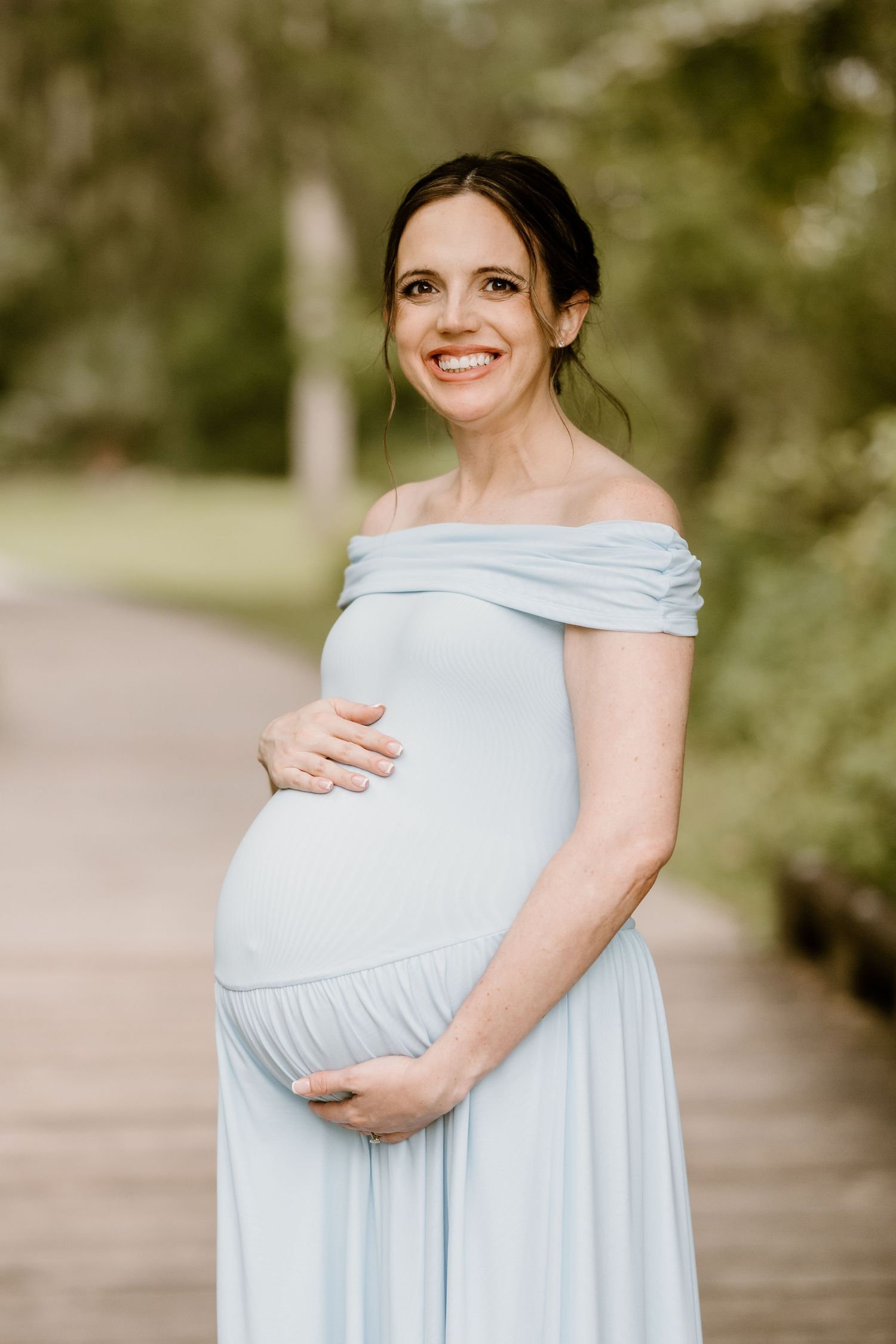 Woman in light blue maternity dress cradling her baby bump outdoors.