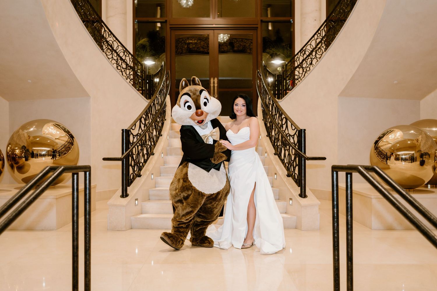 Disney mascots and wedding party pose on elegant staircase with ornate railings.
