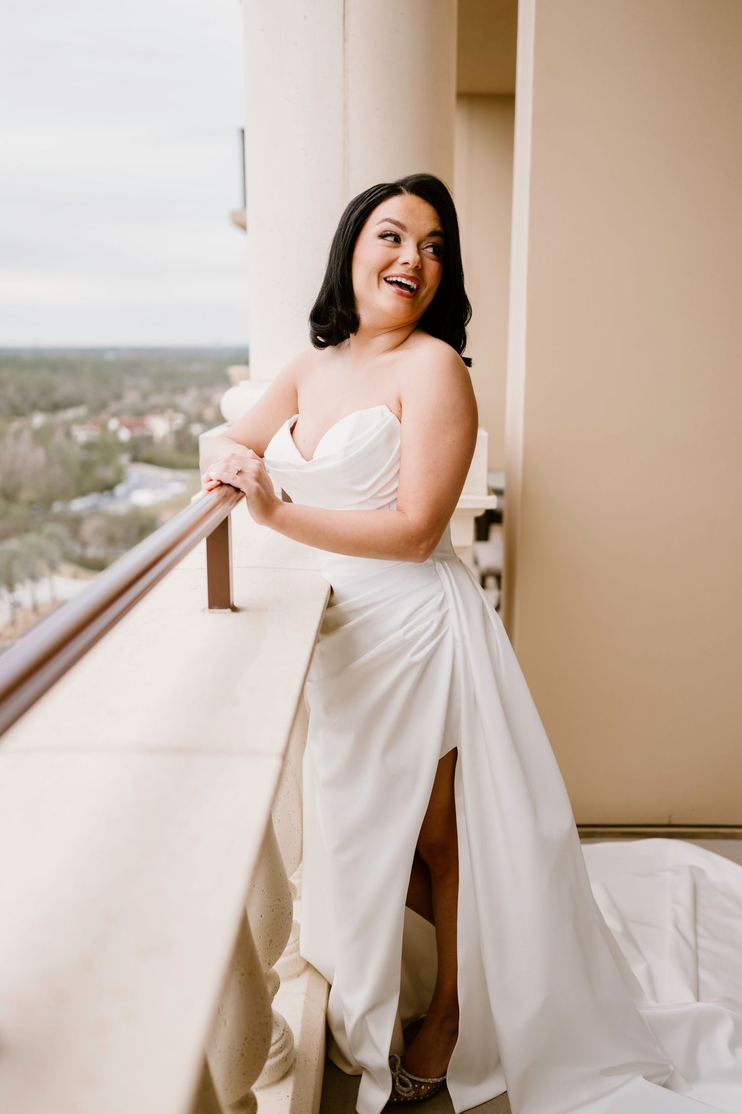 Bride in white dress poses elegantly on balcony with city view.