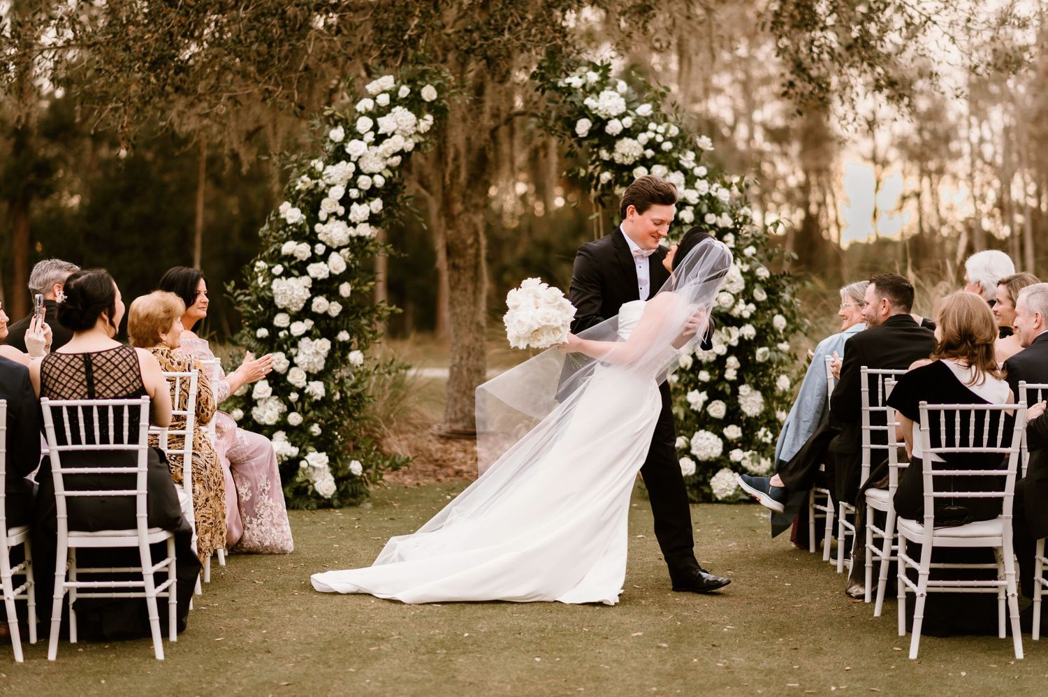 White chairs arranged in rows for an outdoor wedding ceremony with trees in the background.