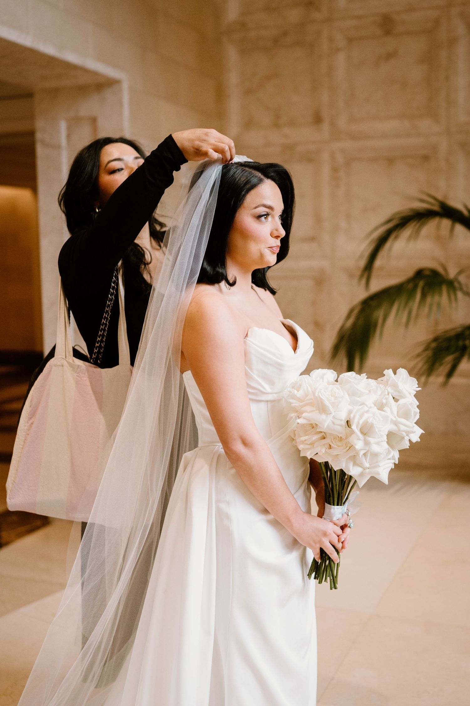 A bride in a white strapless gown holds a bouquet while having her veil adjusted in an elegant indoor setting.