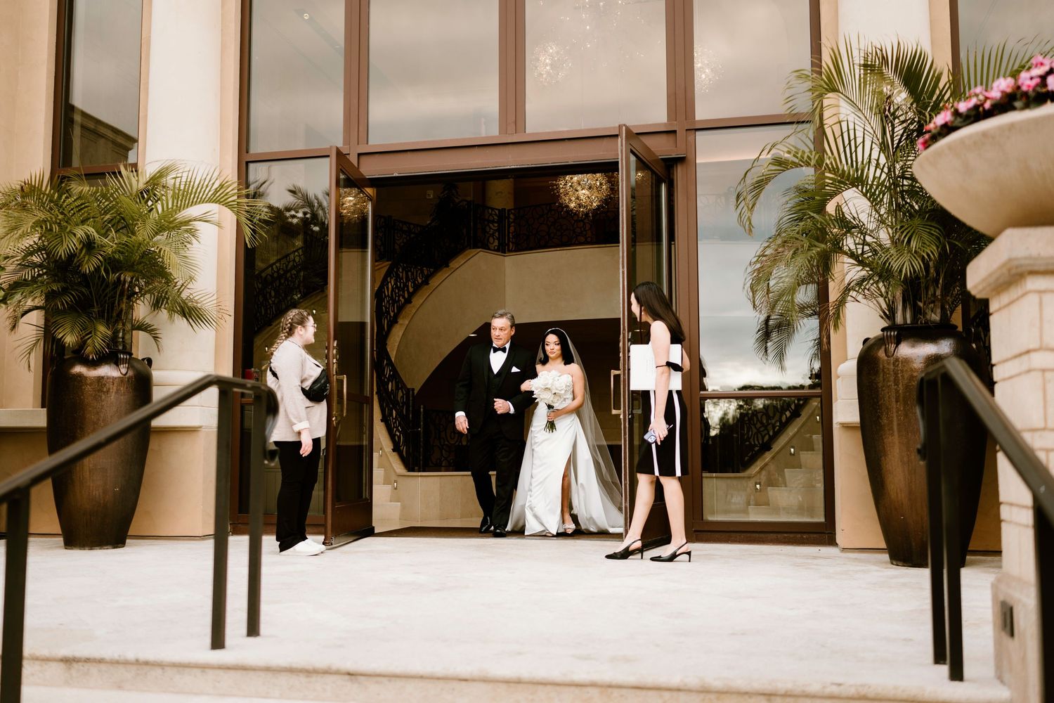 Wedding party exits through glass doors of an elegant building with palm trees and stone columns.