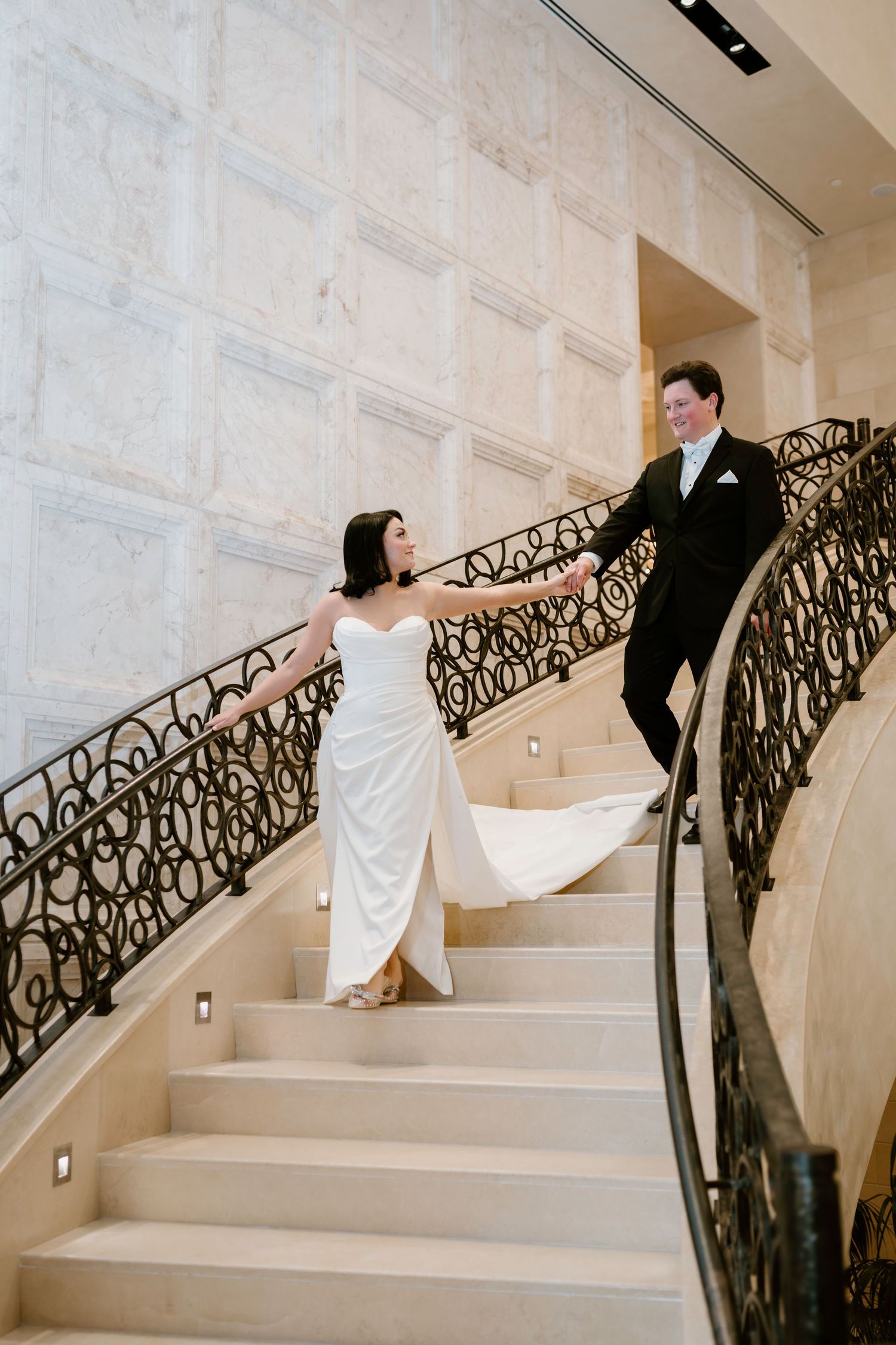 Bride and groom pose on elegant marble staircase with ornate iron railing.