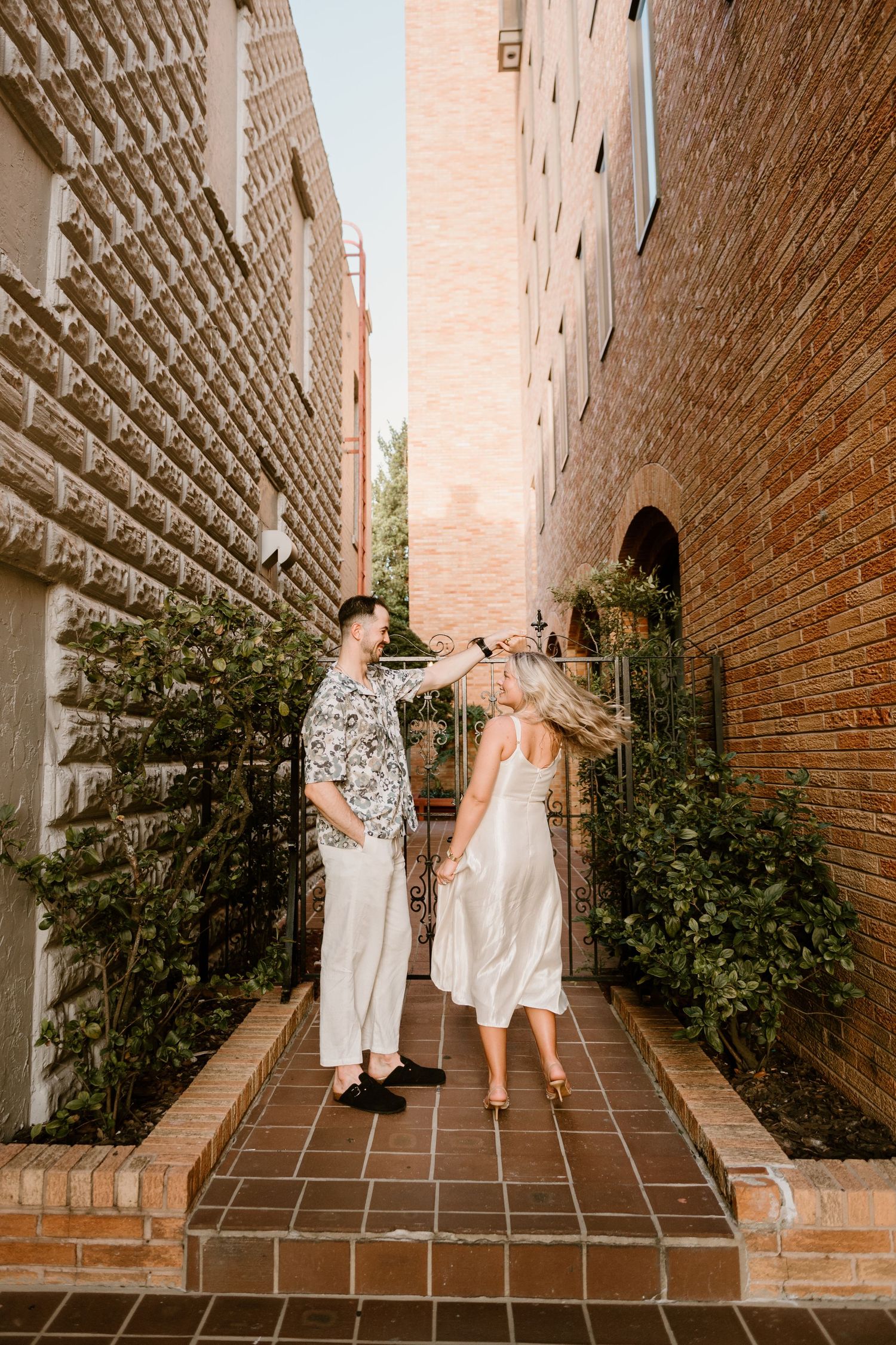 A couple shares a playful moment in a brick-lined outdoor corridor with terracotta tiles and greenery.