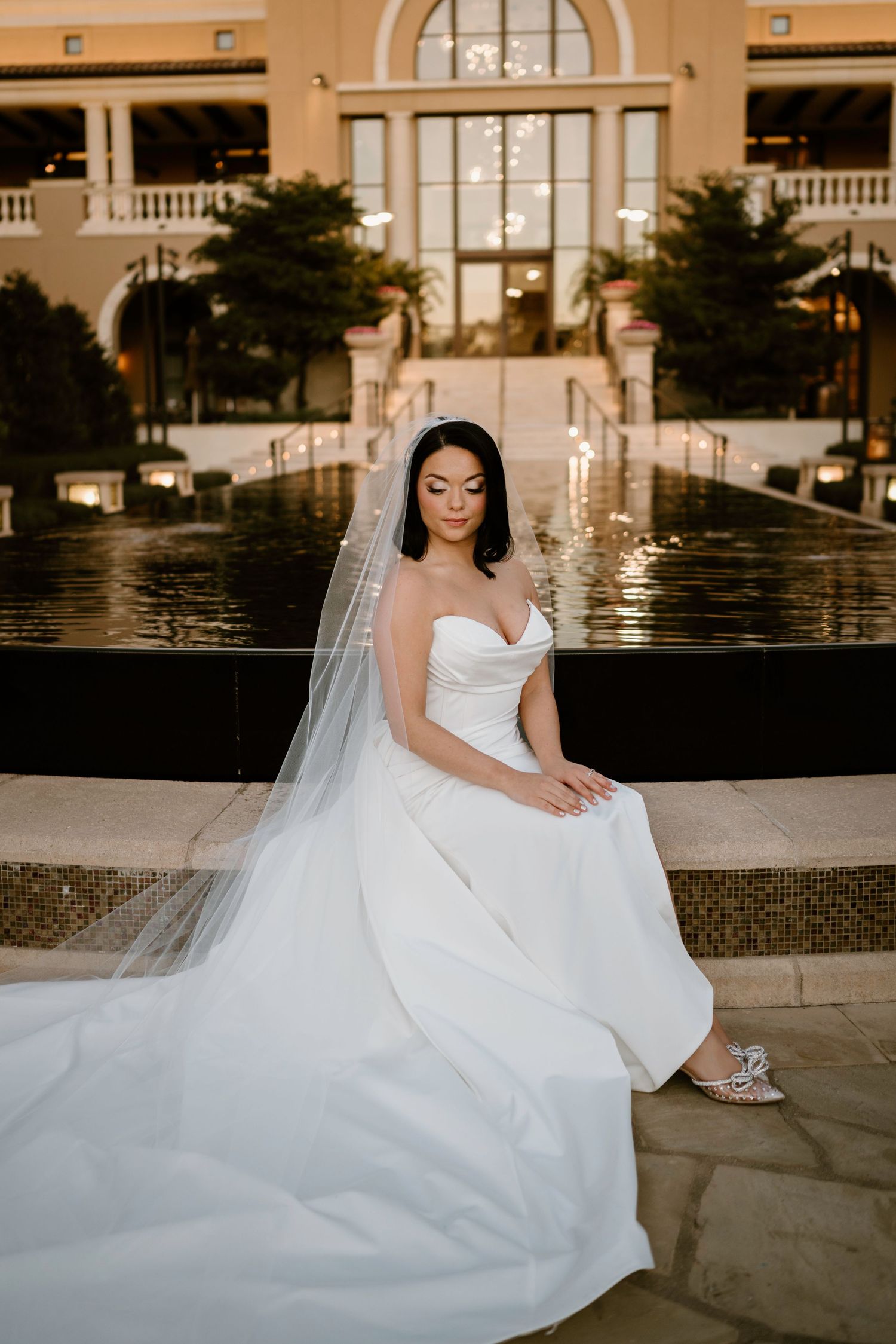 Elegant bride in flowing white gown poses by reflective pool at luxury venue.