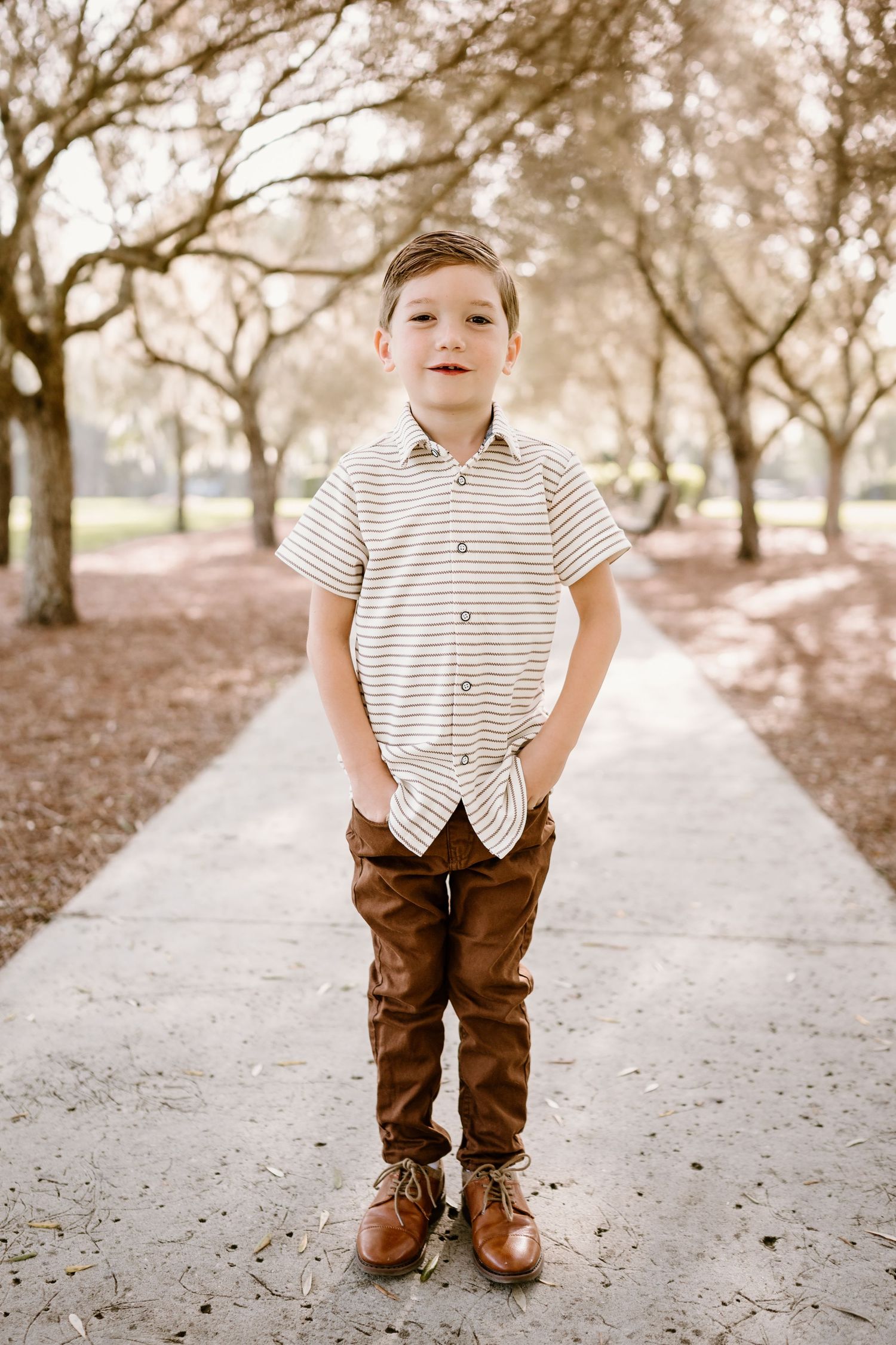 Young boy in striped shirt standing on concrete path surrounded by trees.