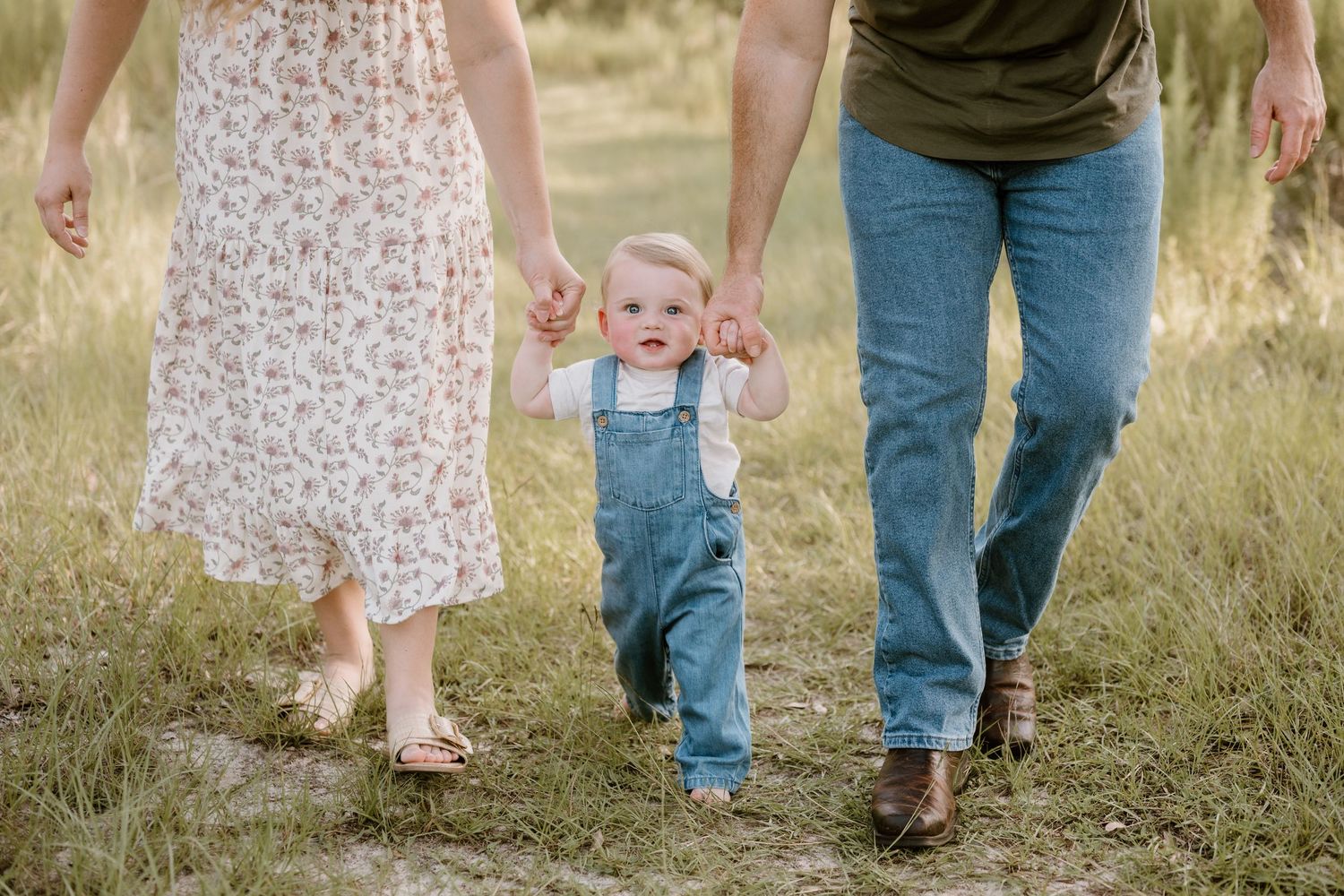 A toddler in denim overalls walks hand-in-hand with parents through tall grass at sunset.