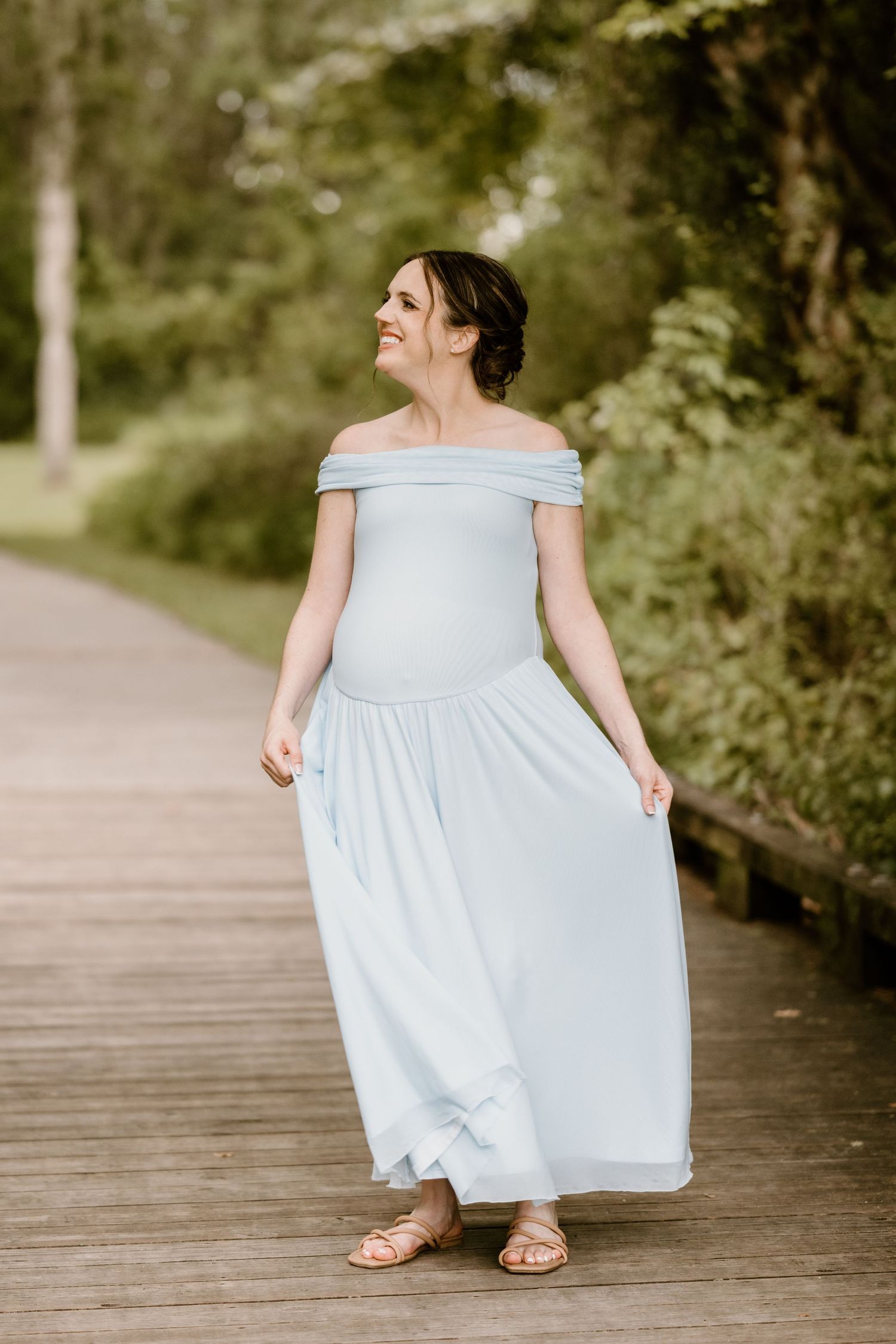 A light blue off-shoulder dress twirls gracefully on a wooden boardwalk surrounded by greenery.