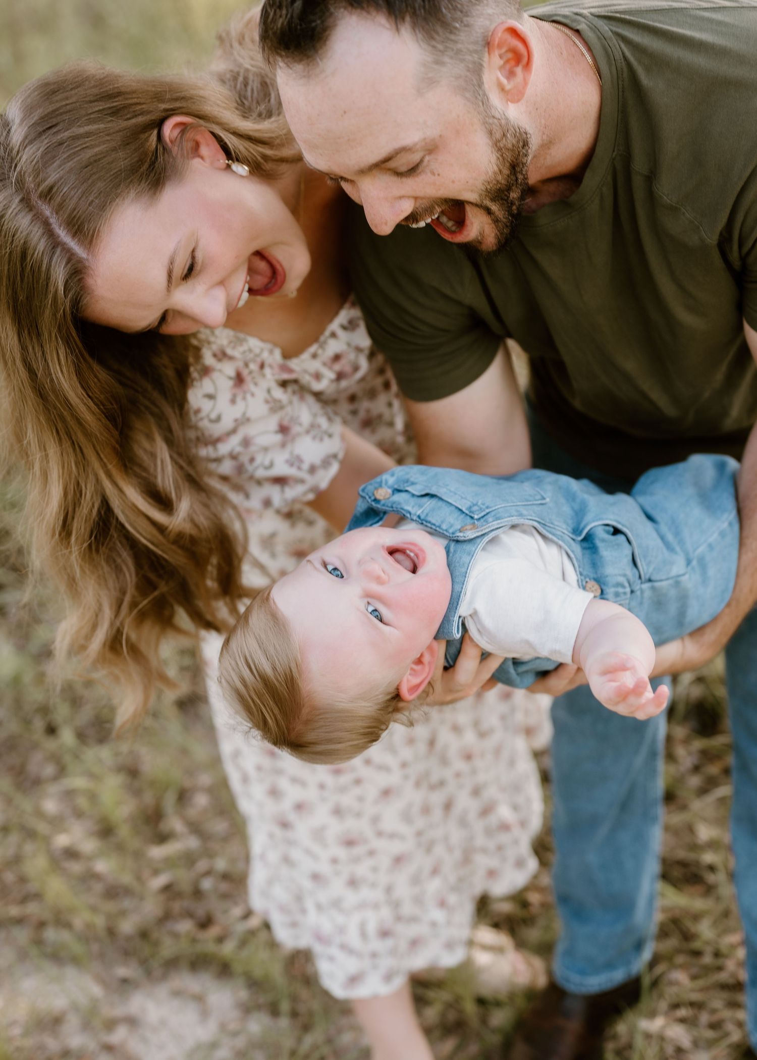 Playful family moment as parents hold their laughing baby upside down in a sunny outdoor setting.