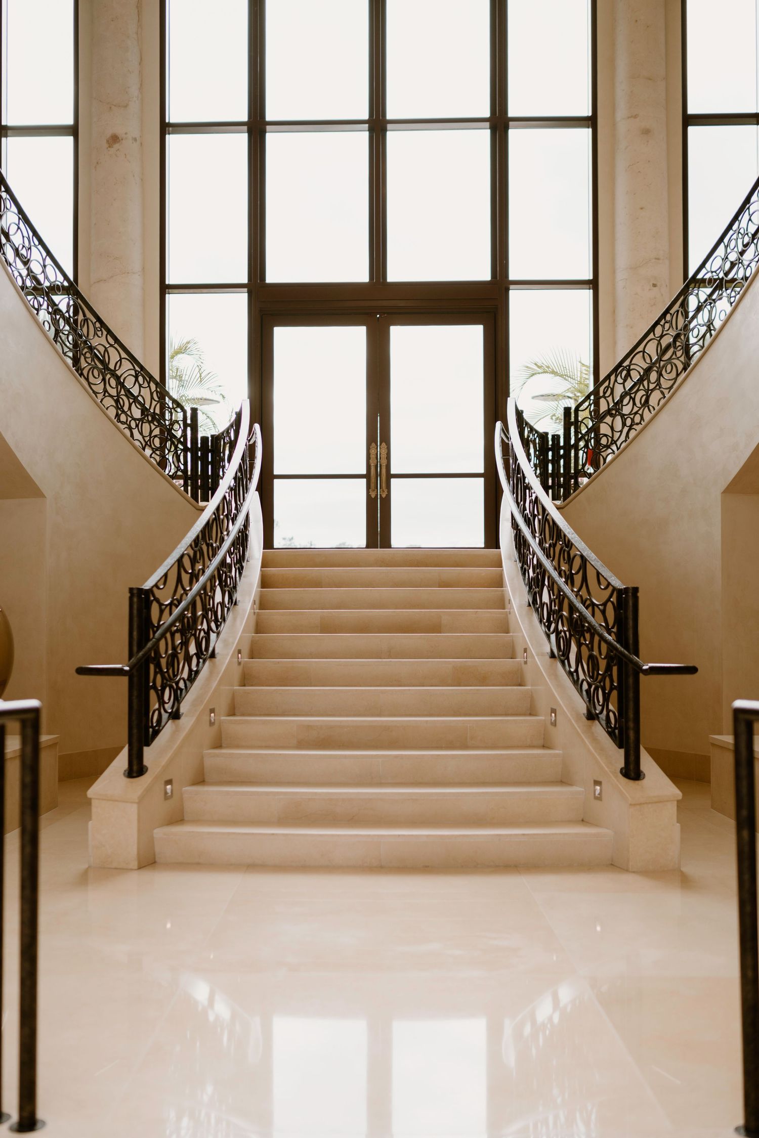 Grand staircase with ornate iron railings and tall windows in an elegant cream-colored interior.