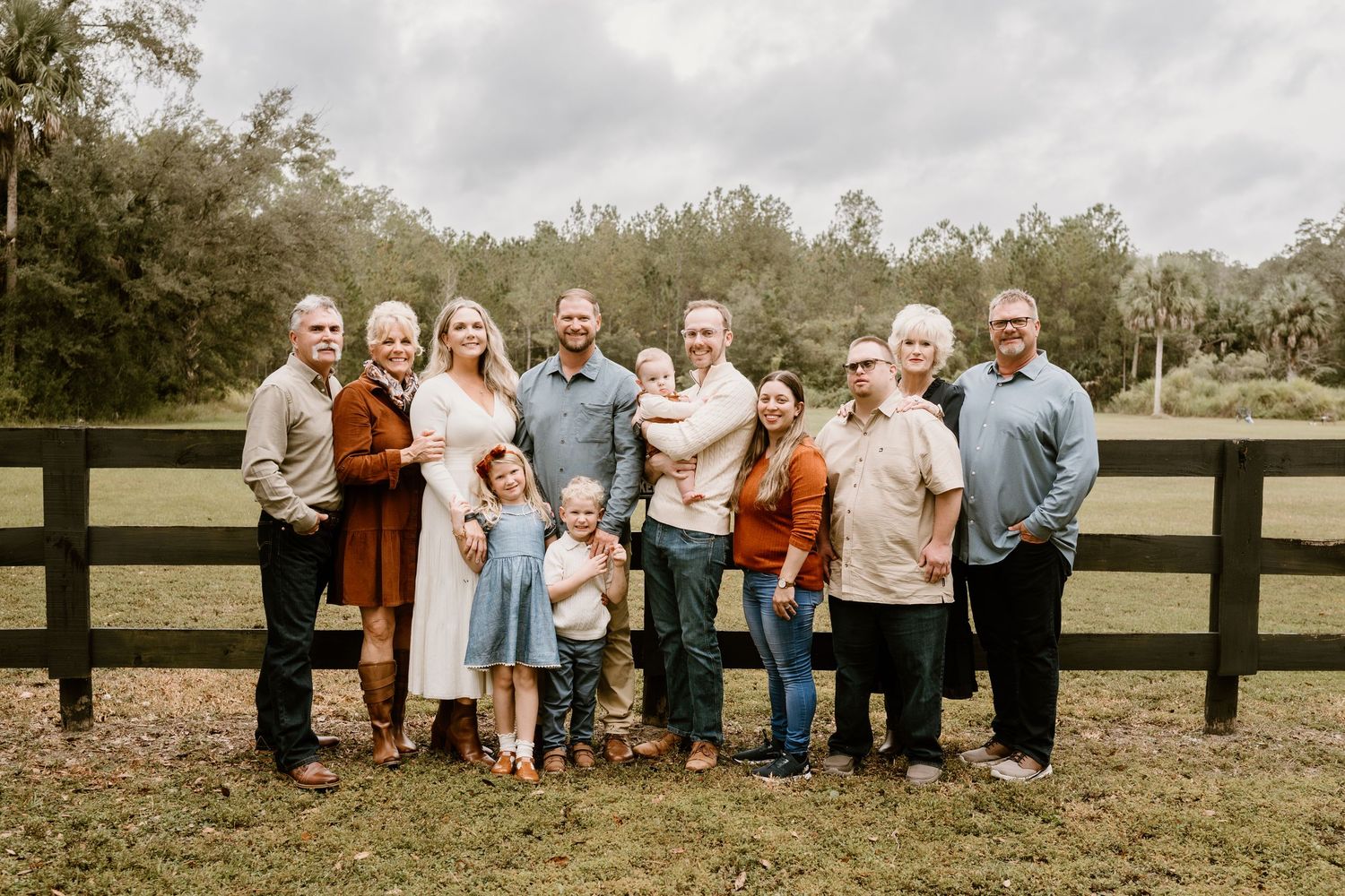 Extended family members pose together on grass near a rustic wooden fence for a group portrait.